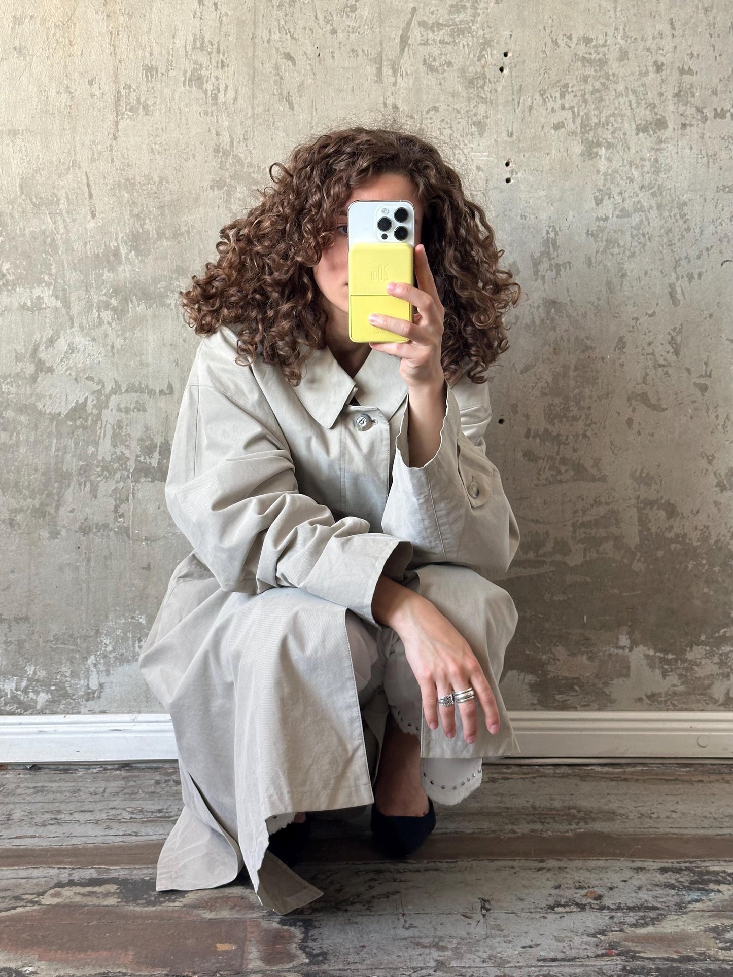 A woman with curly hair is taking a selfie with her phone, holding it up to her face against a concrete wall.