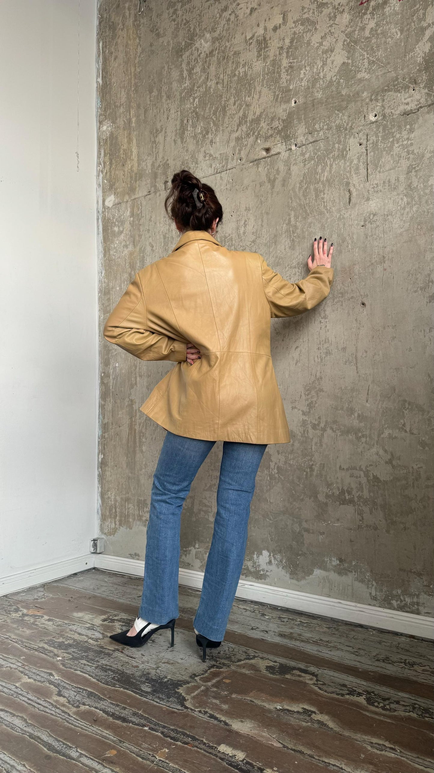 A person wearing a tan jacket and blue jeans stands in front of a concrete wall, with their back to the camera.