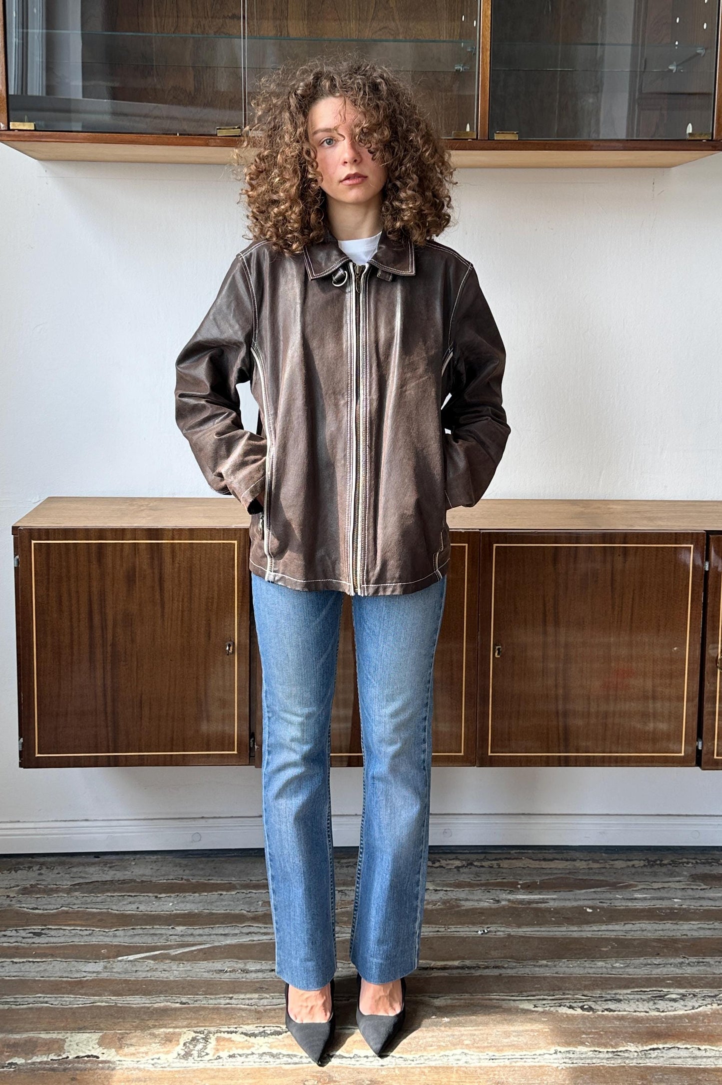 A woman with curly hair stands in a room, wearing a brown leather jacket and blue jeans.