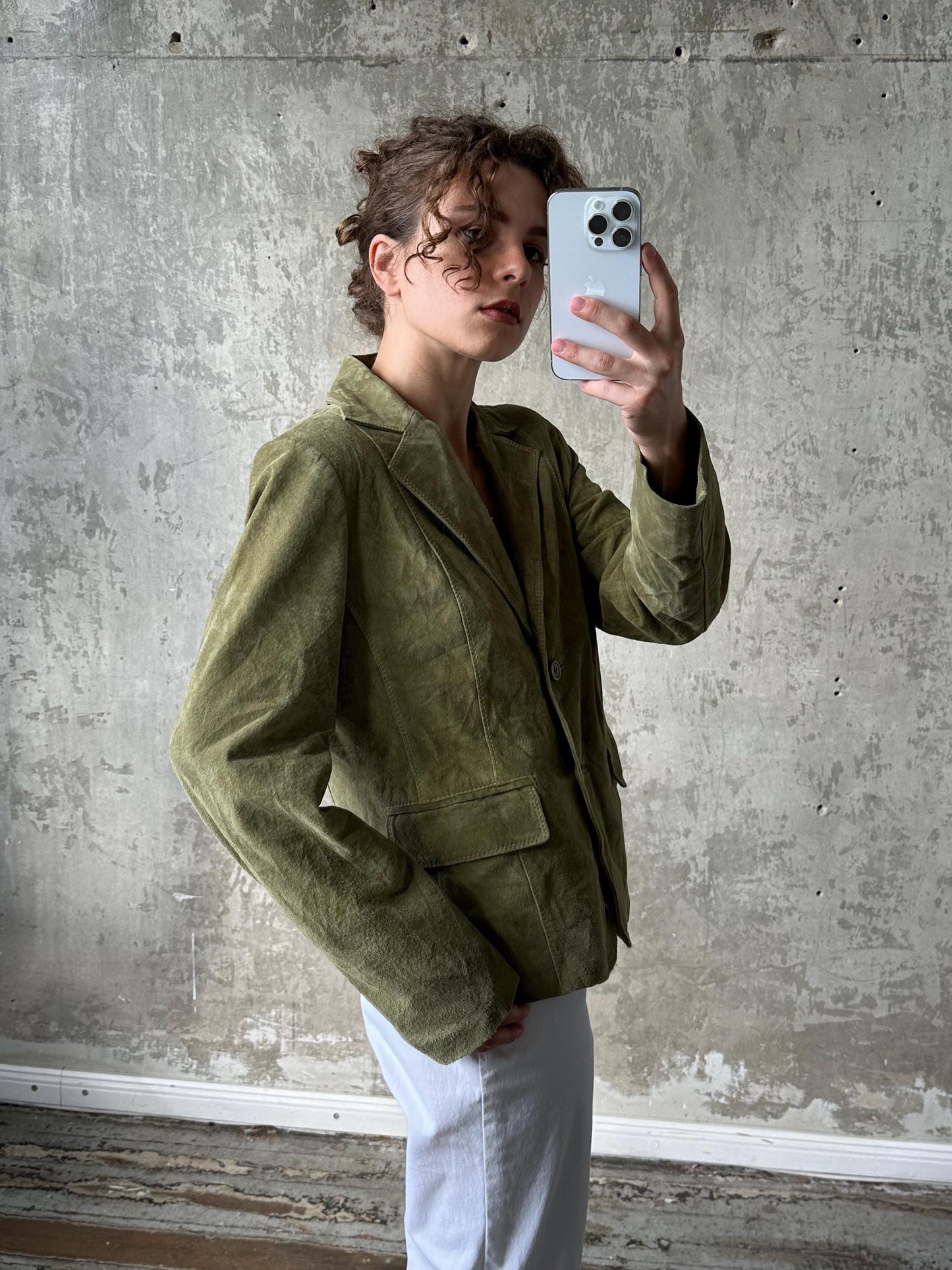 A person is taking a selfie with a white iPhone, wearing a green blazer, and standing in front of a concrete wall.