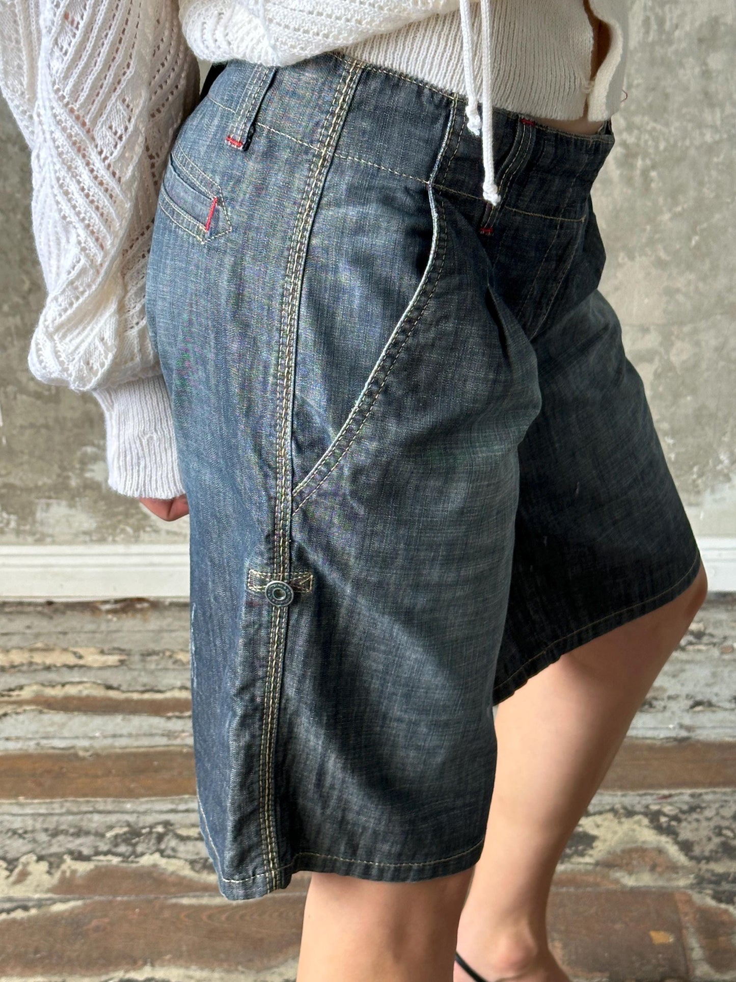 A person wearing a white sweater and blue jeans stands in front of a concrete wall.