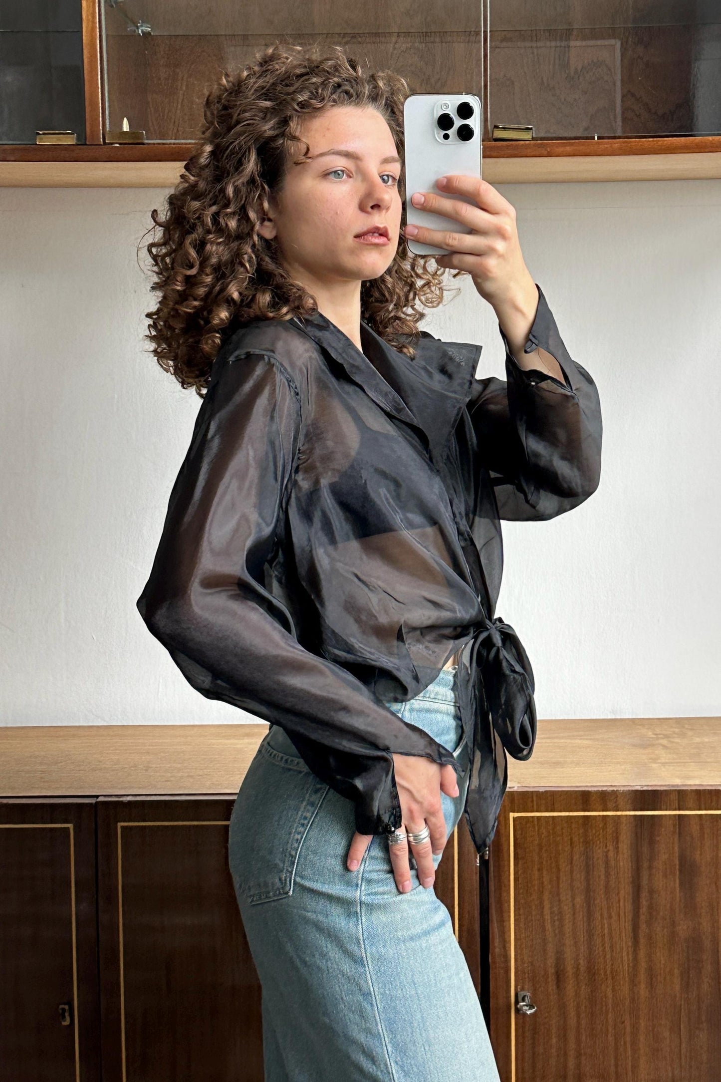 A woman with curly hair is taking a selfie with a white iPhone in front of a wooden cabinet.
