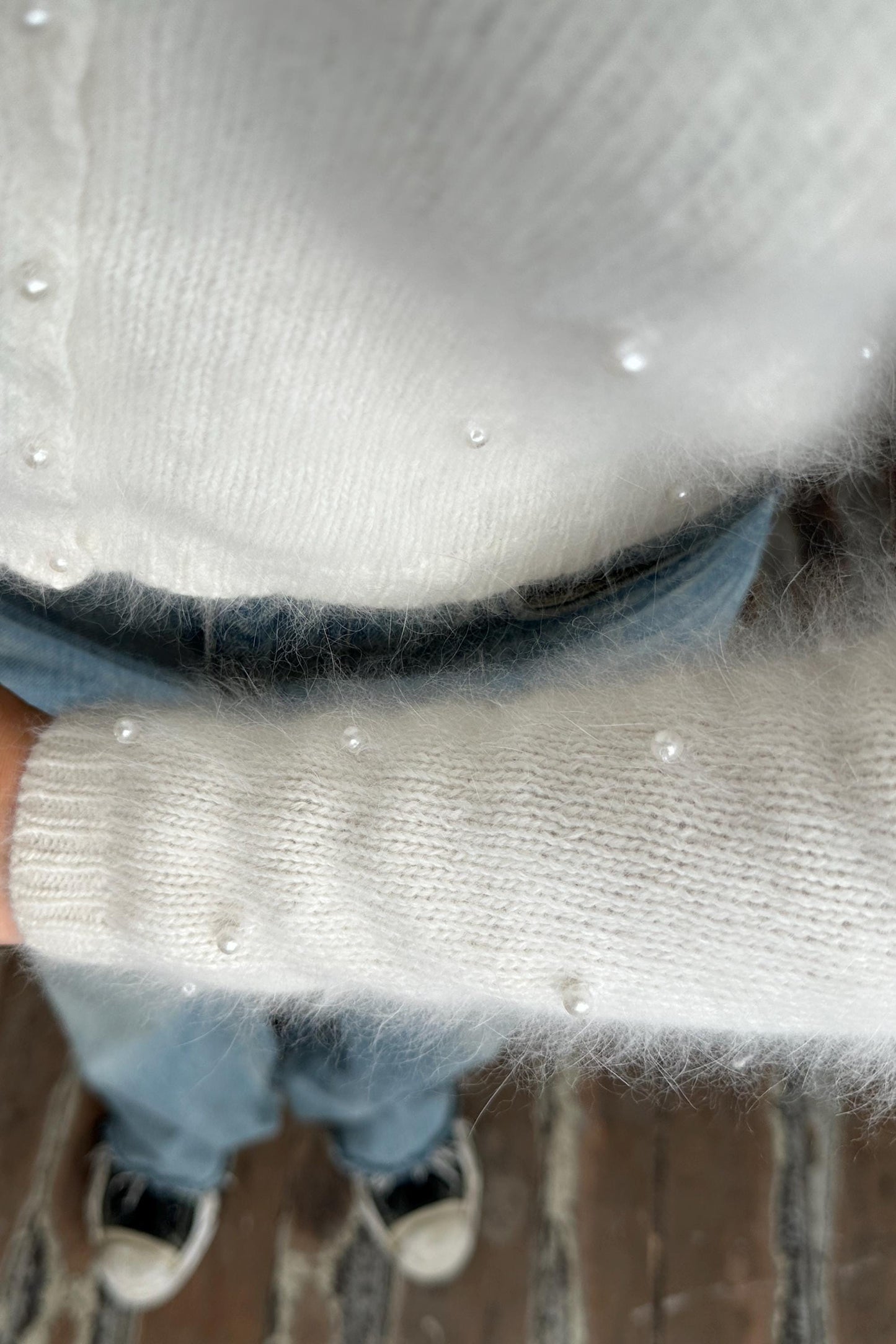 A close-up view of a person wearing a white sweater with small white pearls on it, standing on a wooden floor.