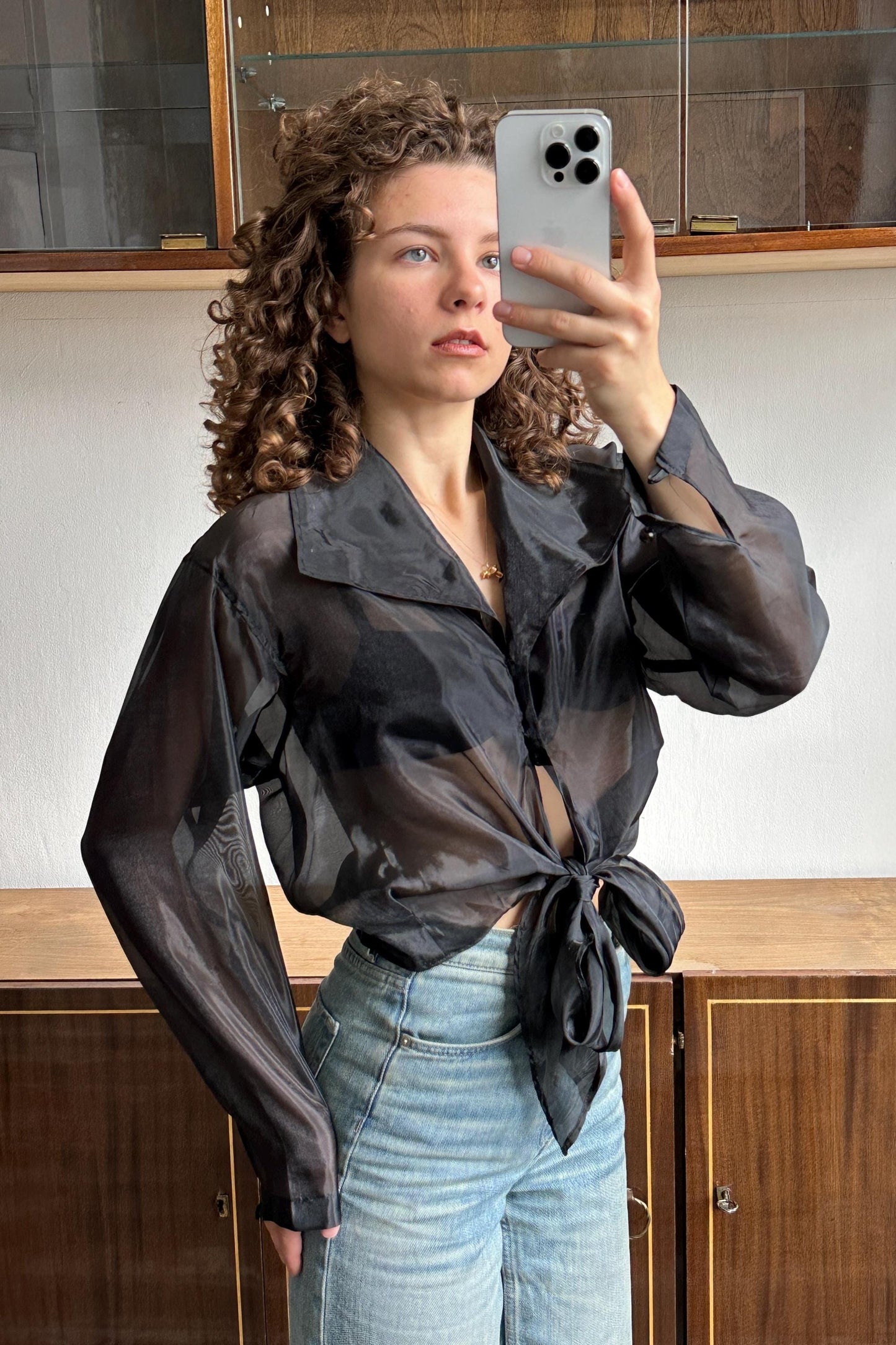 A woman with curly hair is taking a selfie with a smartphone, wearing a black blouse and blue jeans.