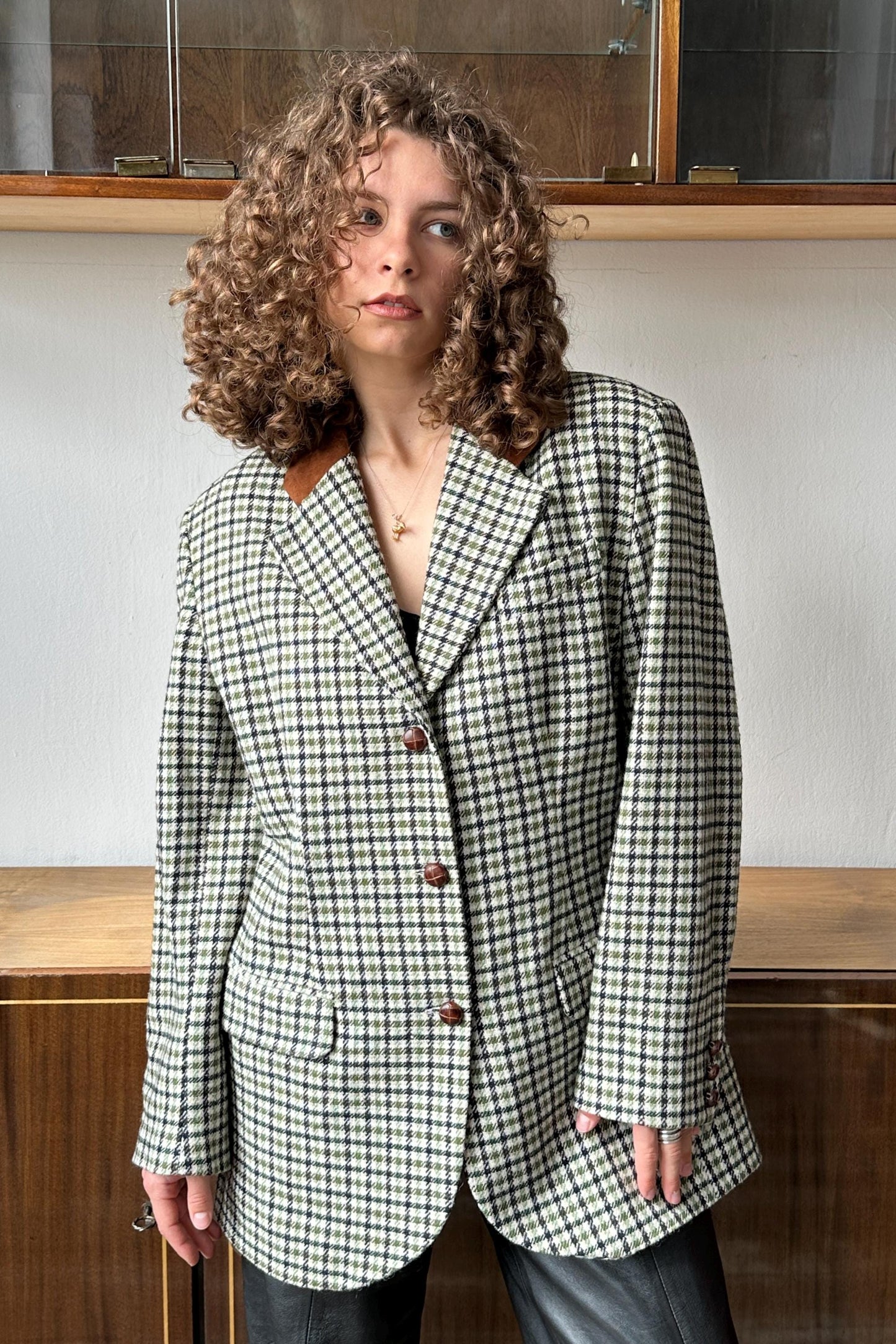 A woman with curly hair wearing a black and white checkered blazer stands in front of a wooden cabinet.