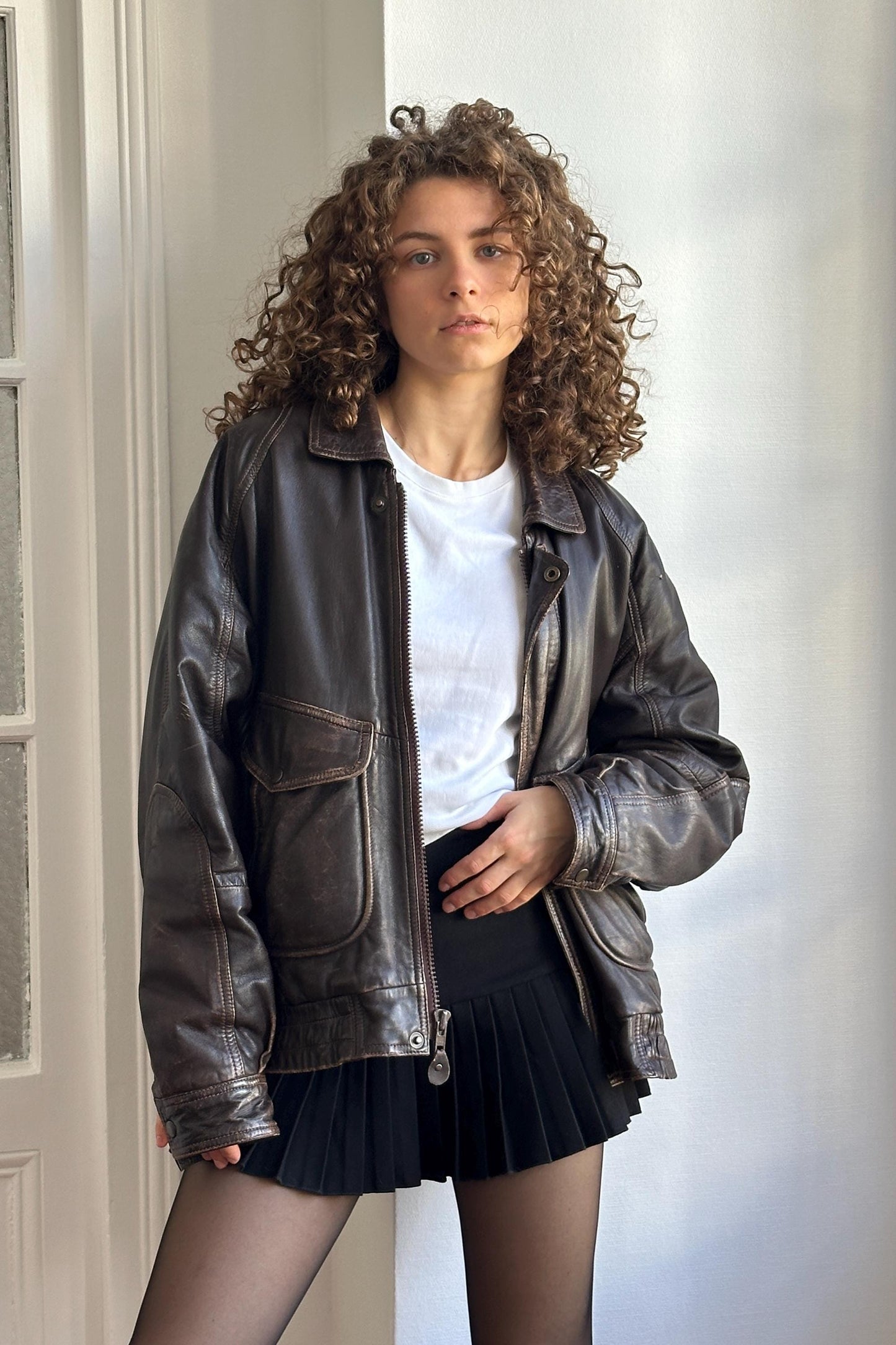 A young woman with curly hair wearing a brown leather jacket and a black skirt stands in front of a white wall.