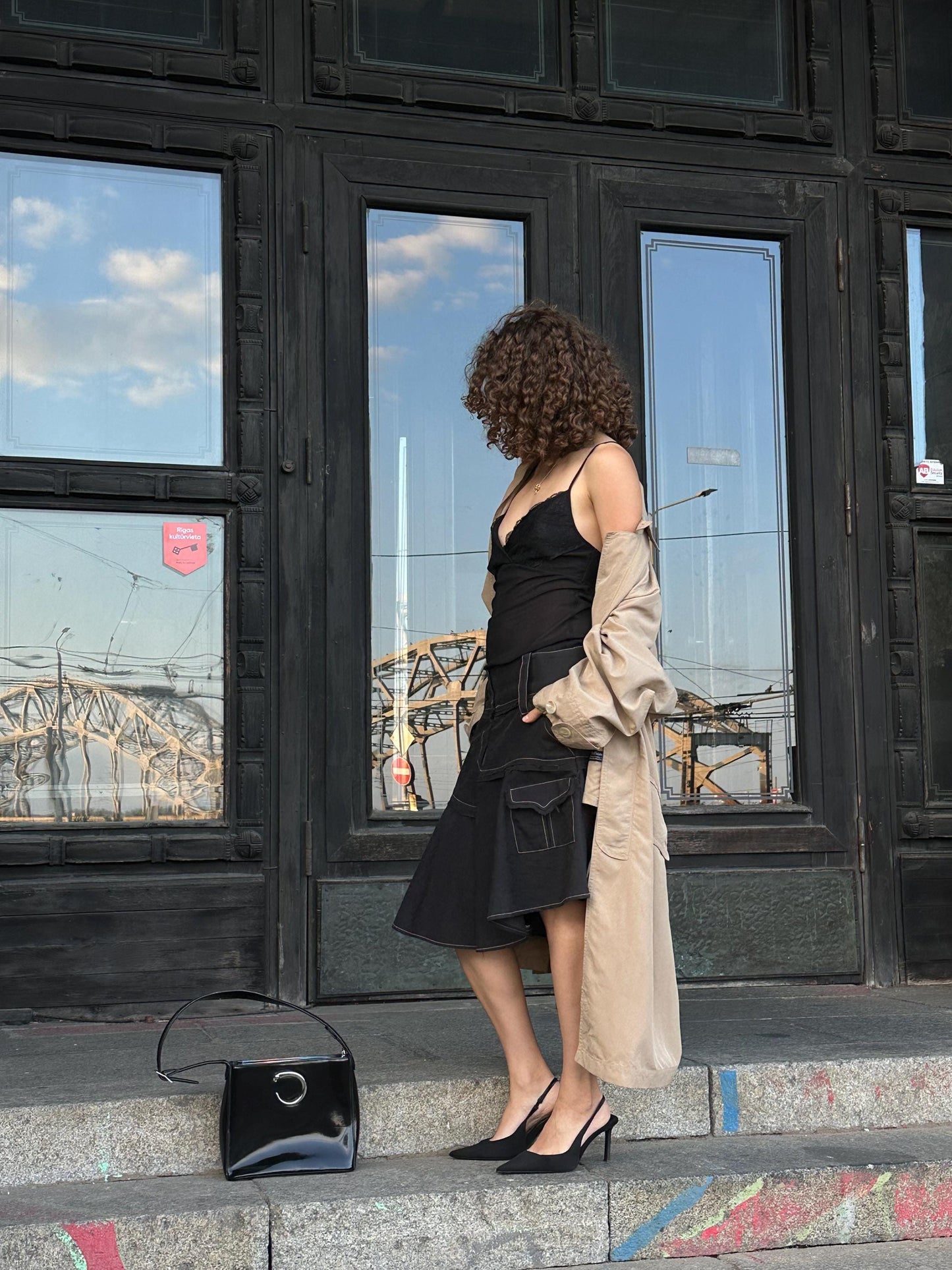 A woman in a black dress and beige coat stands on a city street, with a black purse and a black handbag on the ground in front of her.