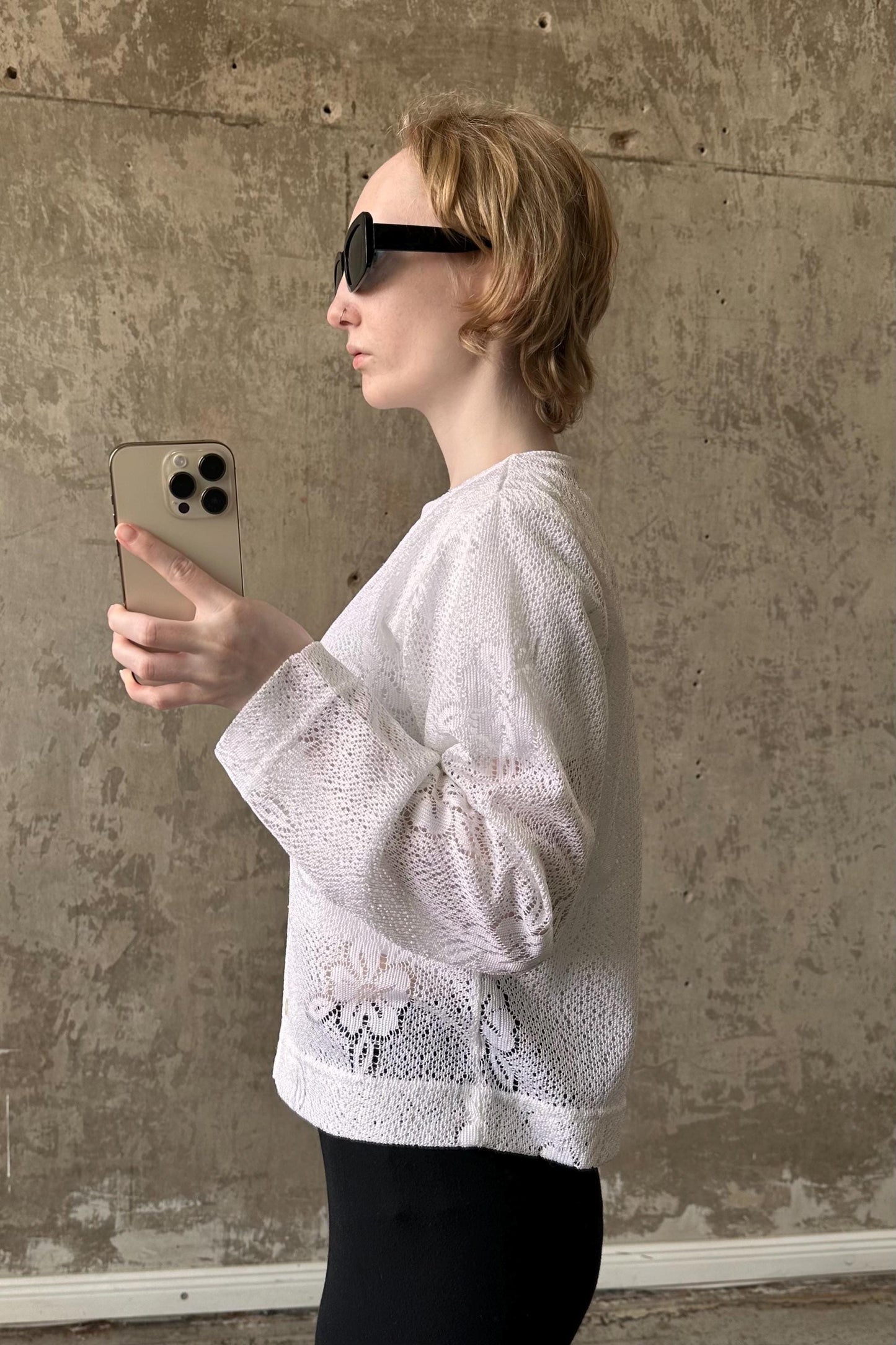 A woman wearing a white lace top and sunglasses is holding a smartphone and looking at it.
