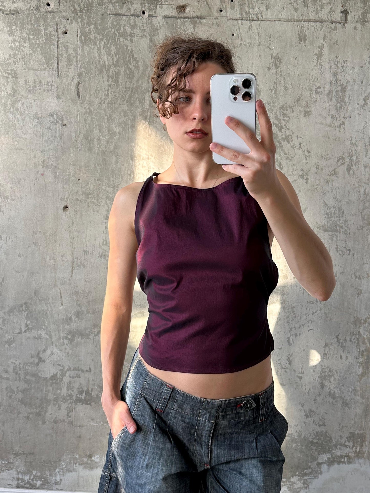 A woman in a purple top is taking a selfie with a smartphone, standing in front of a concrete wall.