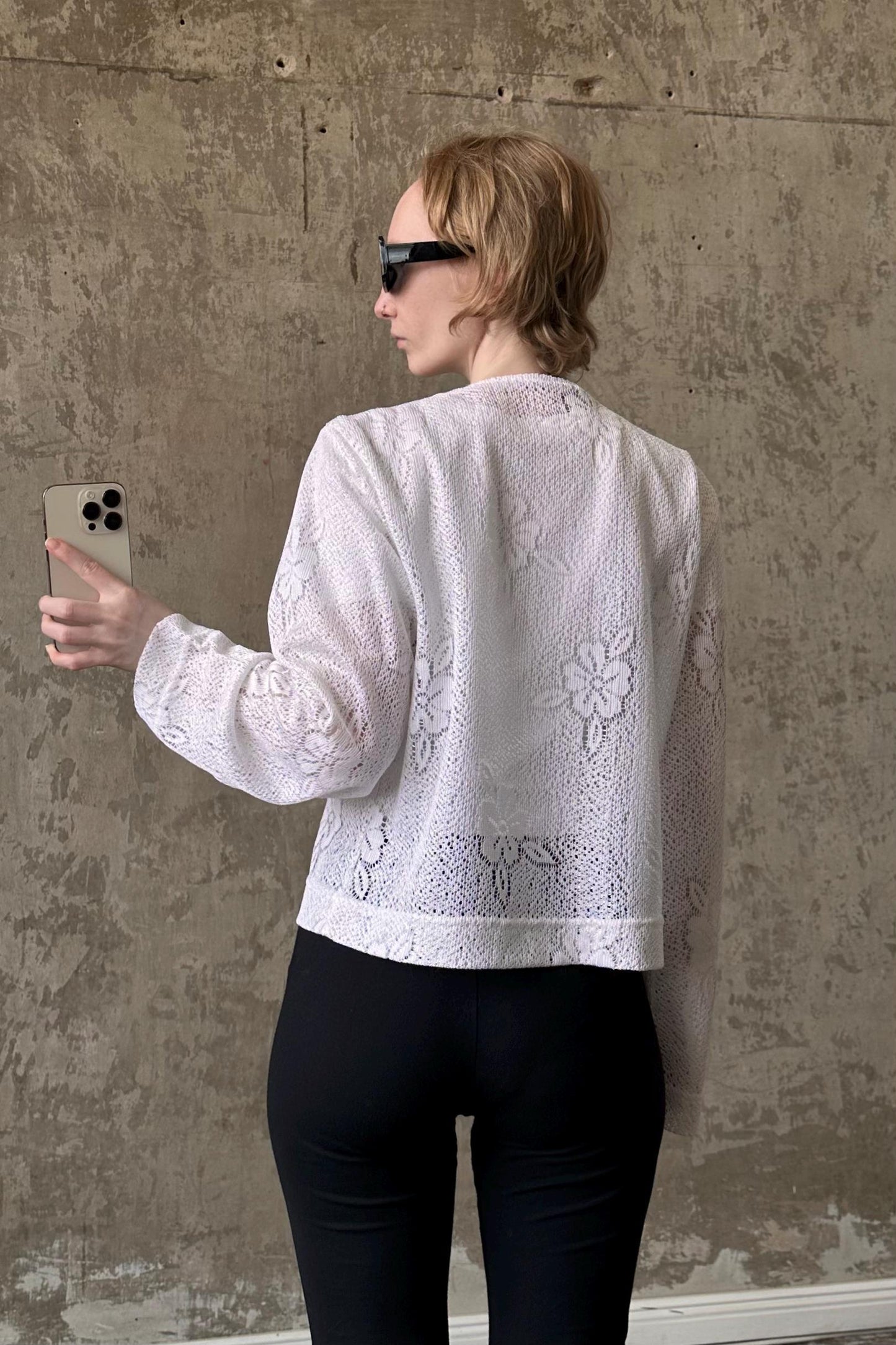 A woman wearing a white lace top and black pants is holding a smartphone and looking at it, with a concrete wall in the background.