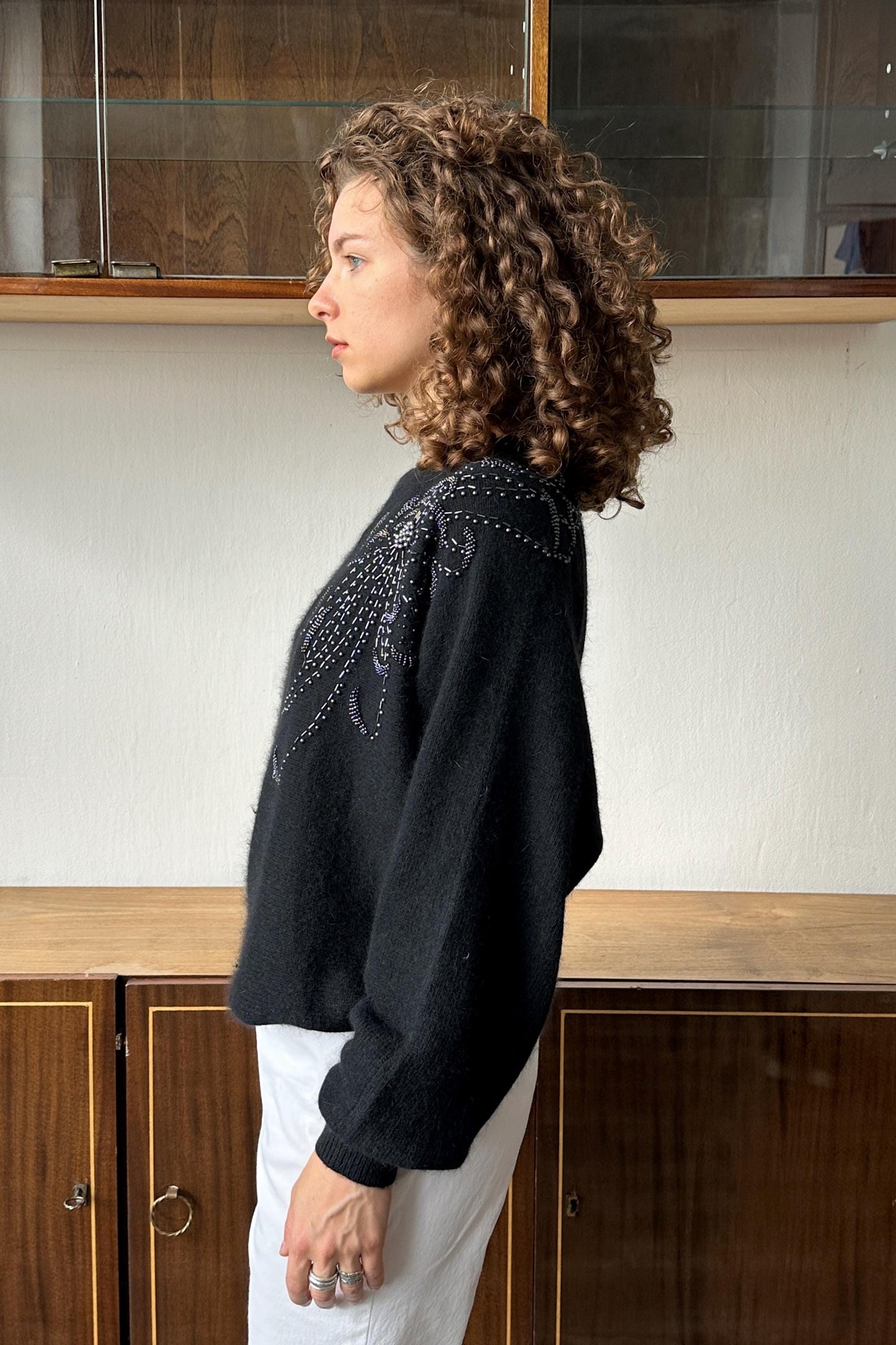 A woman with curly hair wearing a black sweater with a floral pattern stands in front of a wooden cabinet.