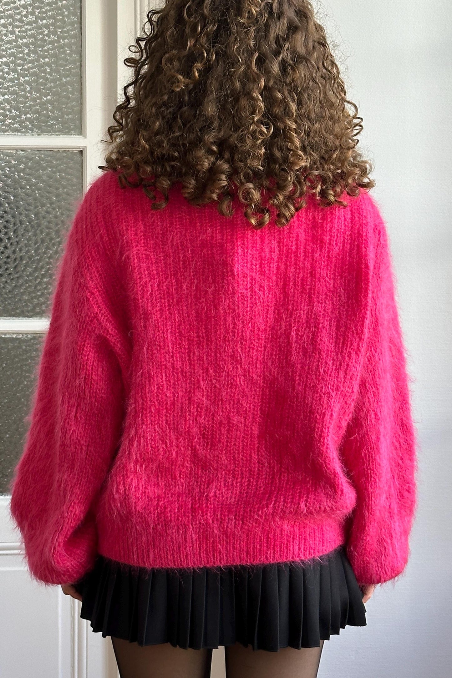 A woman with curly hair wearing a bright pink sweater and a black skirt stands in front of a glass door.