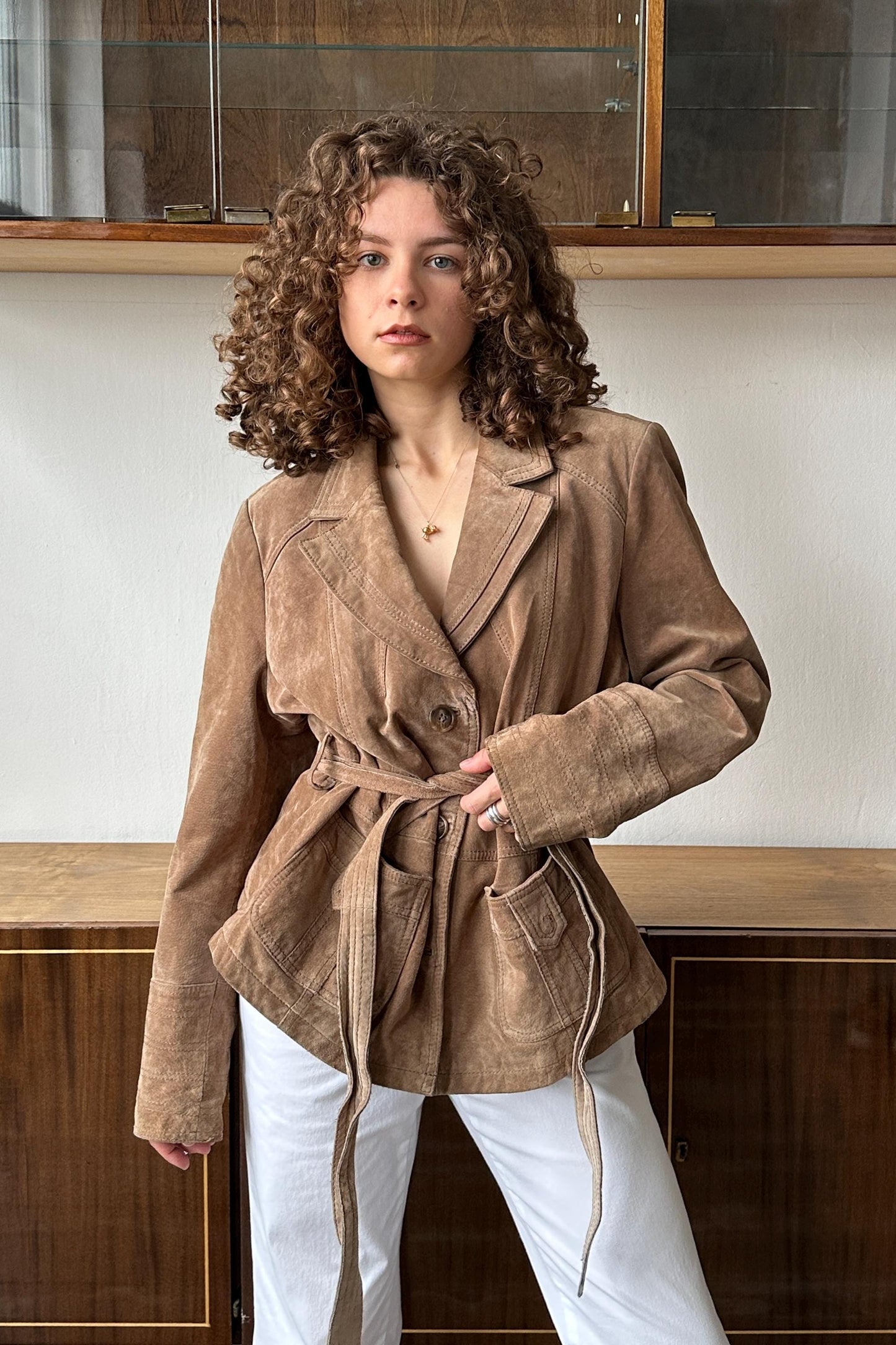 A woman with curly hair wearing a tan jacket stands in front of a wooden cabinet.