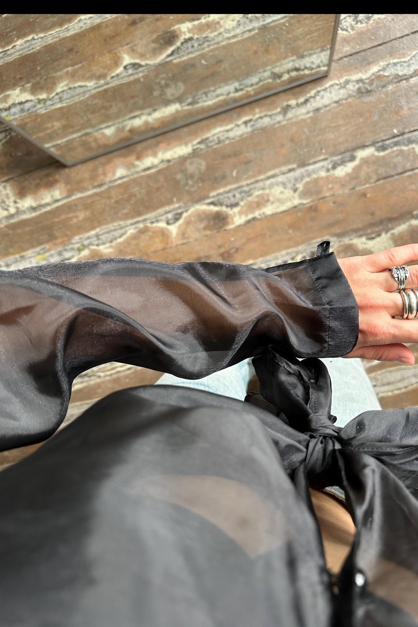 A close-up view of a person's arm wearing a black sheer blouse, with a ring visible on their finger.