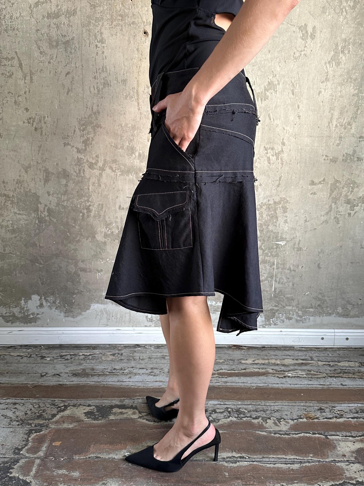 A person wearing a black skirt with pockets and high heels stands in front of a concrete wall.