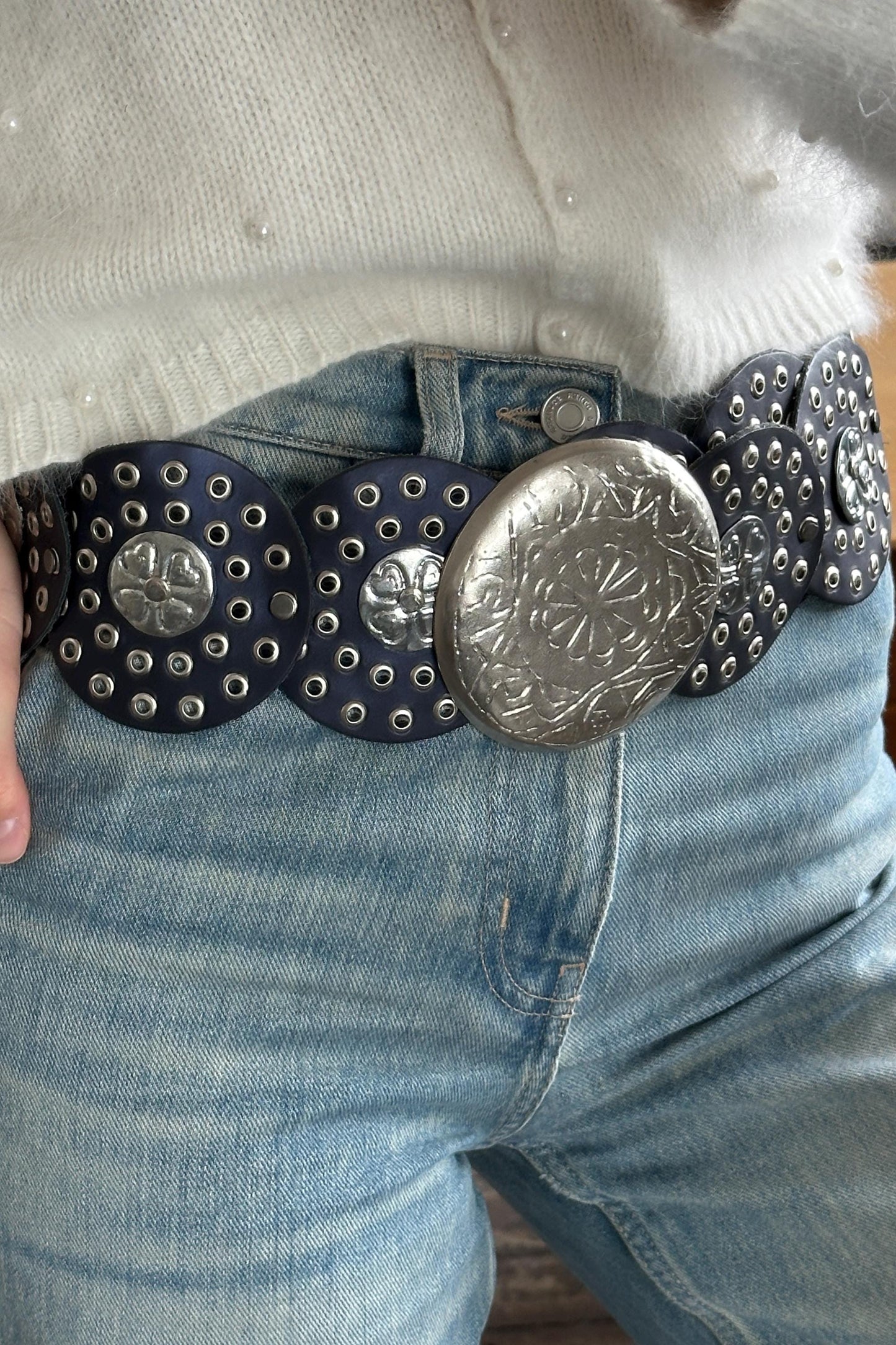 A close-up view of a person wearing a white sweater and blue jeans, with a large, decorative belt featuring a metal buckle and circular studs.