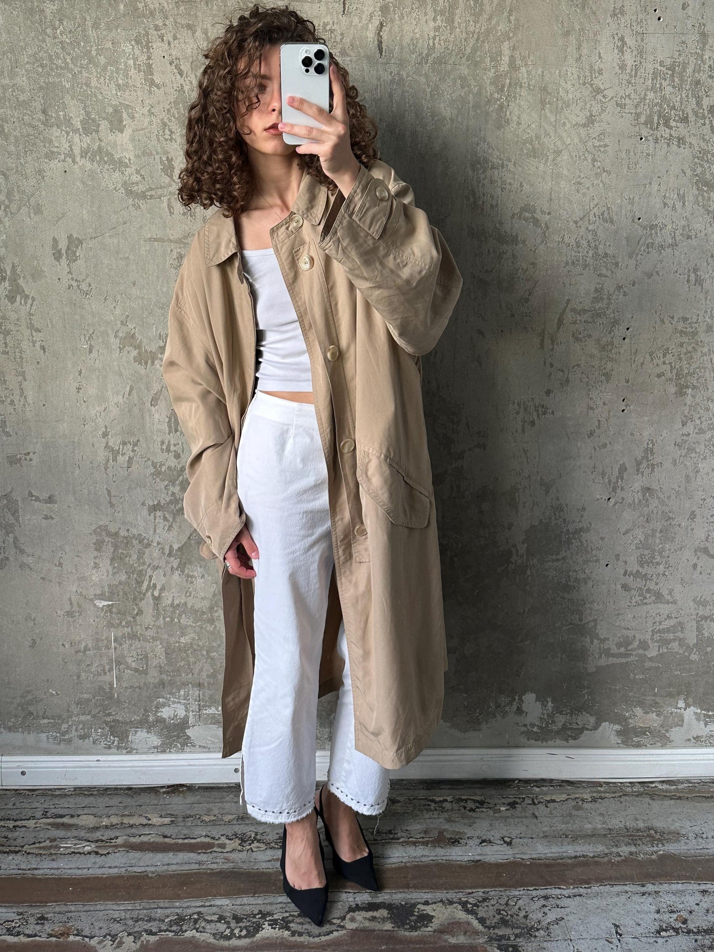 A woman with curly hair is taking a selfie with her phone, wearing a beige coat and white pants against a concrete wall.