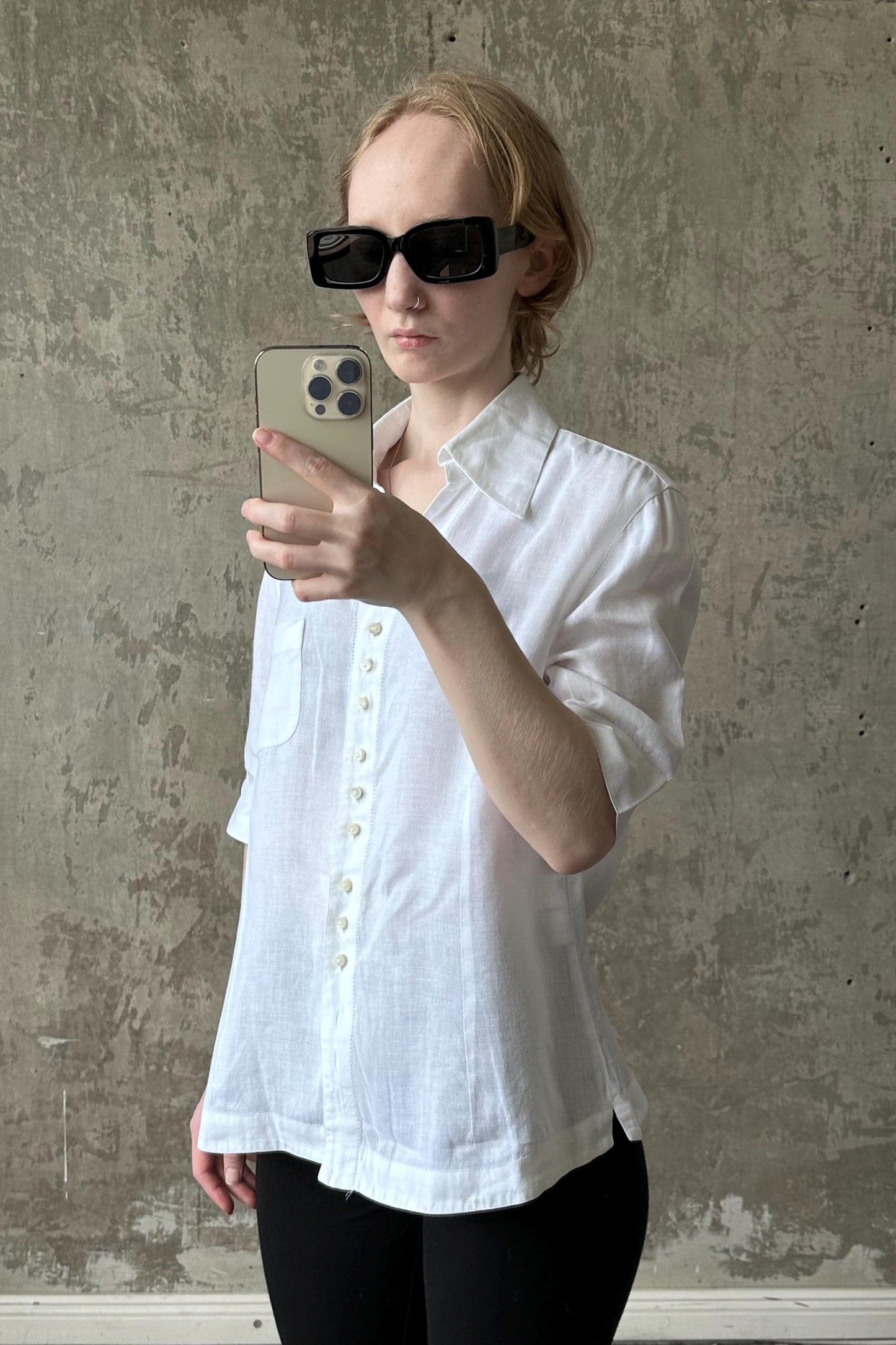 A woman wearing a white shirt and sunglasses is taking a selfie with her iPhone against a concrete wall.