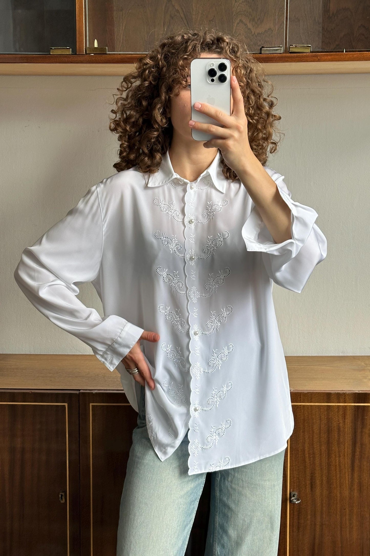 A woman with curly hair is taking a selfie with her iPhone, wearing a white blouse with embroidered details.