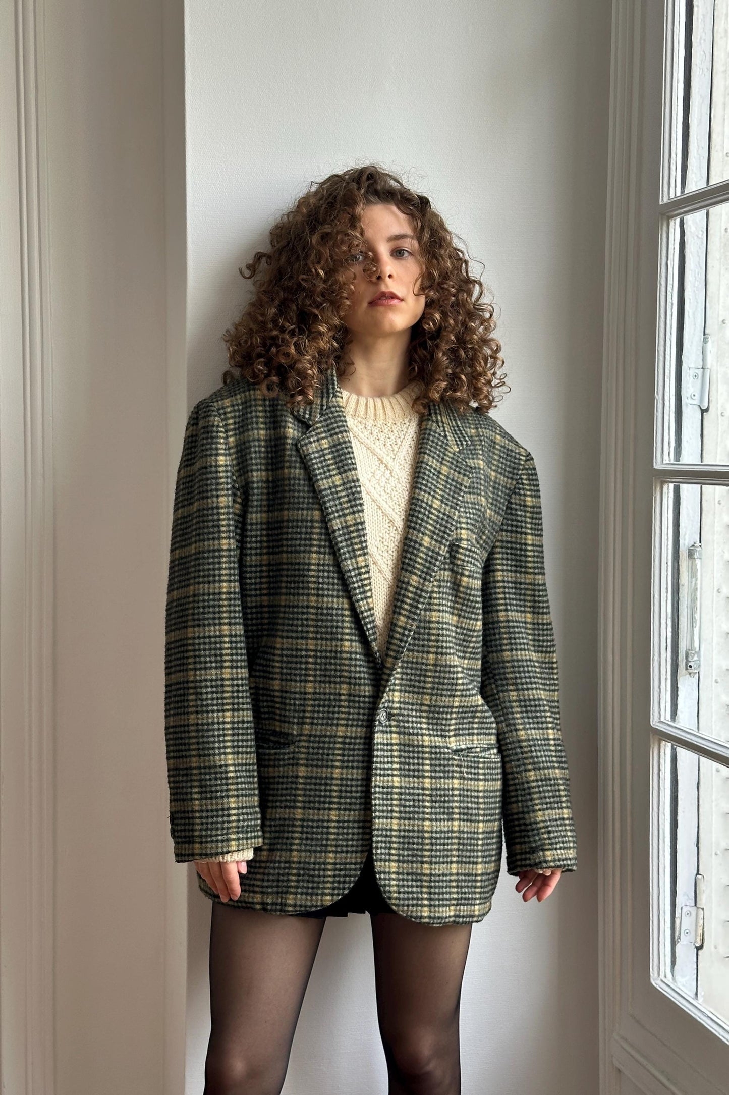 A woman with curly hair wearing a plaid jacket and standing in front of a window.