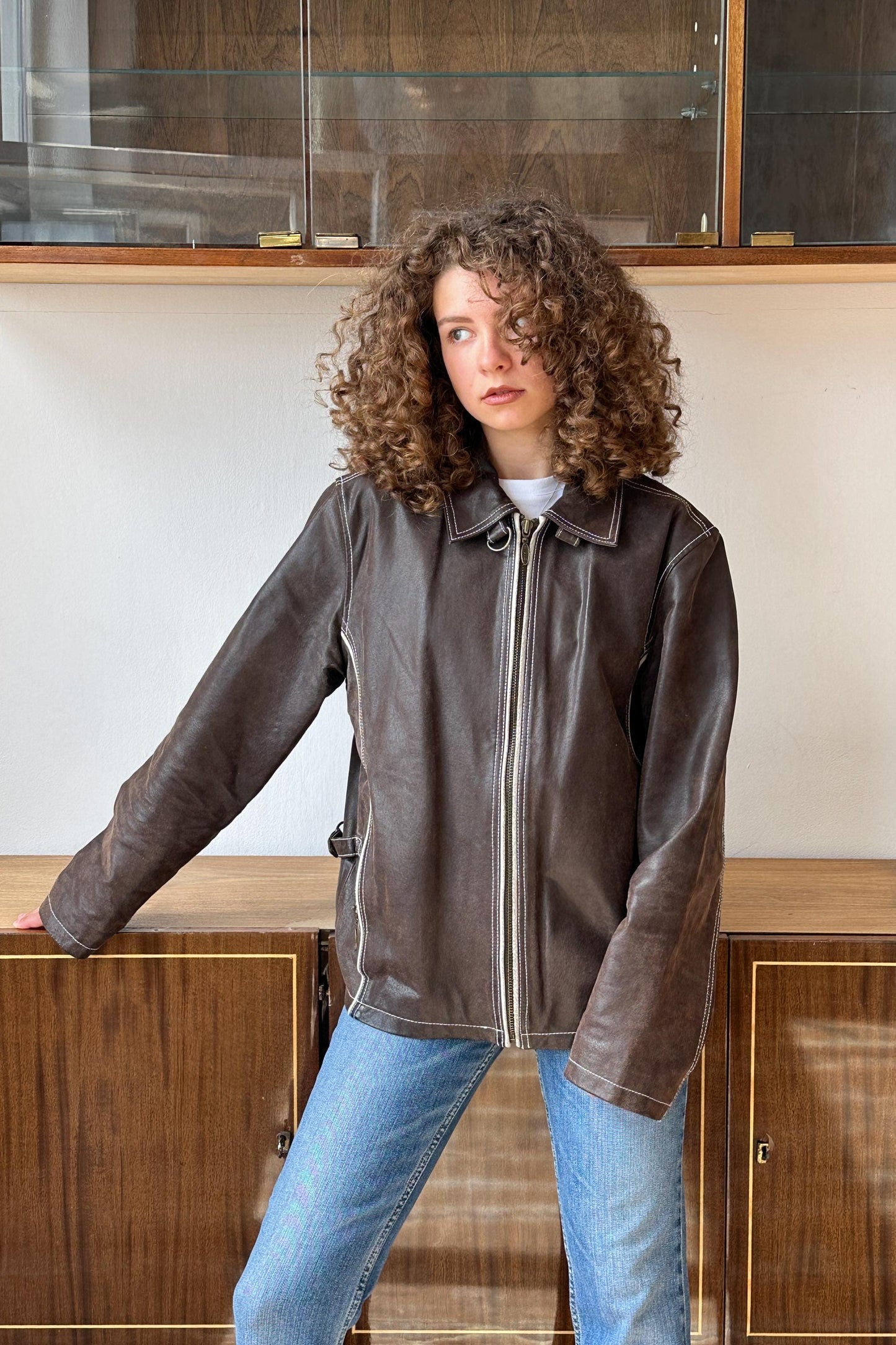 A young woman with curly hair wearing a brown leather jacket and blue jeans stands in front of a wooden cabinet.