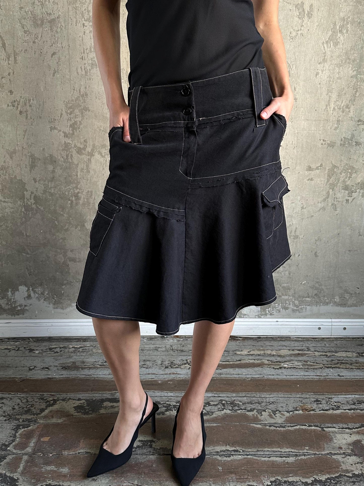 A person wearing a black skirt with pockets and high heels stands in front of a concrete wall.