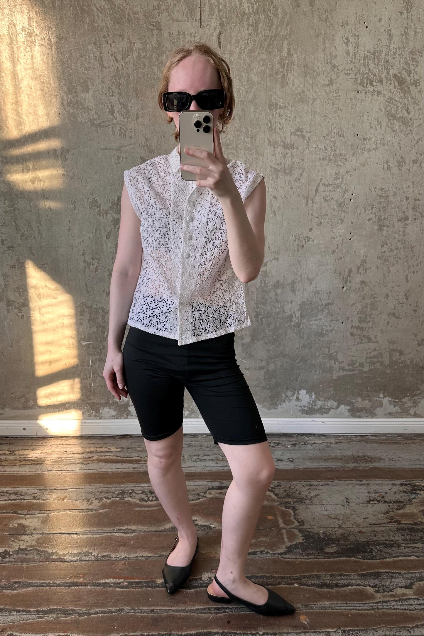 A person is taking a selfie with a smartphone, wearing a white lace top, black shorts, and black shoes, standing in front of a concrete wall.