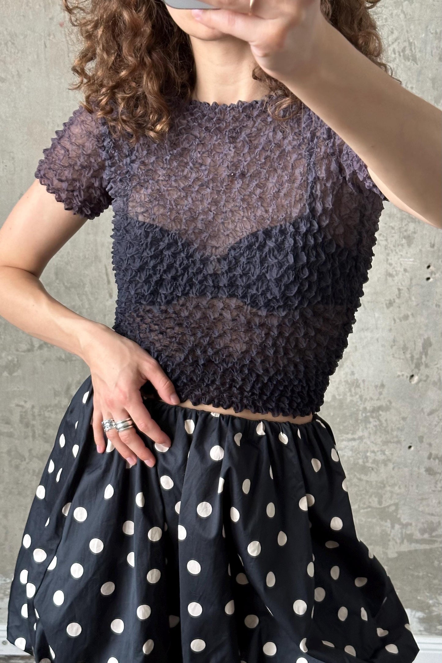 A woman wearing a black and white polka dot skirt and a gray ruffled top, posing with her hand on her hip.