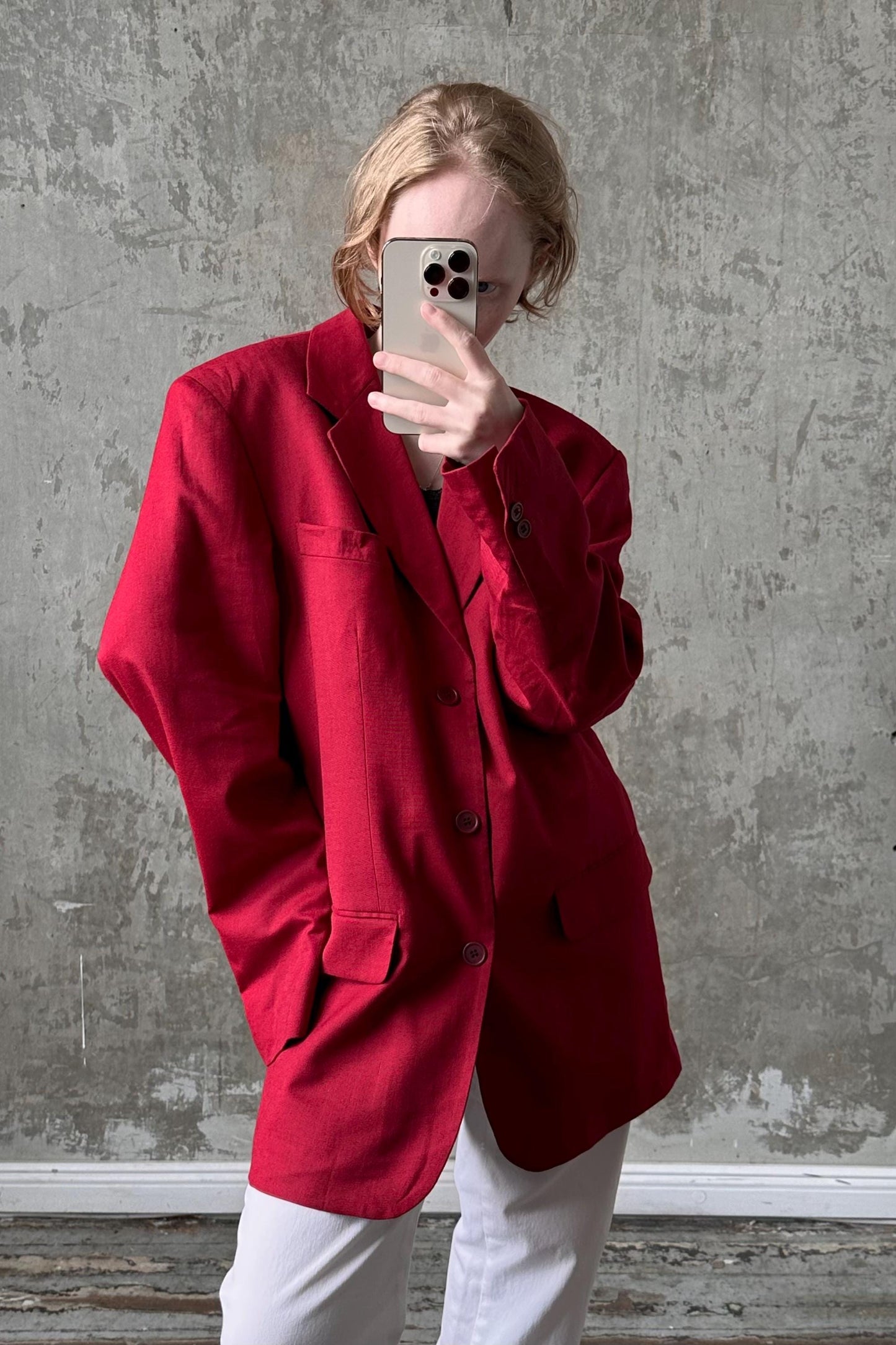 A person wearing a red blazer is taking a selfie with a smartphone against a concrete wall.