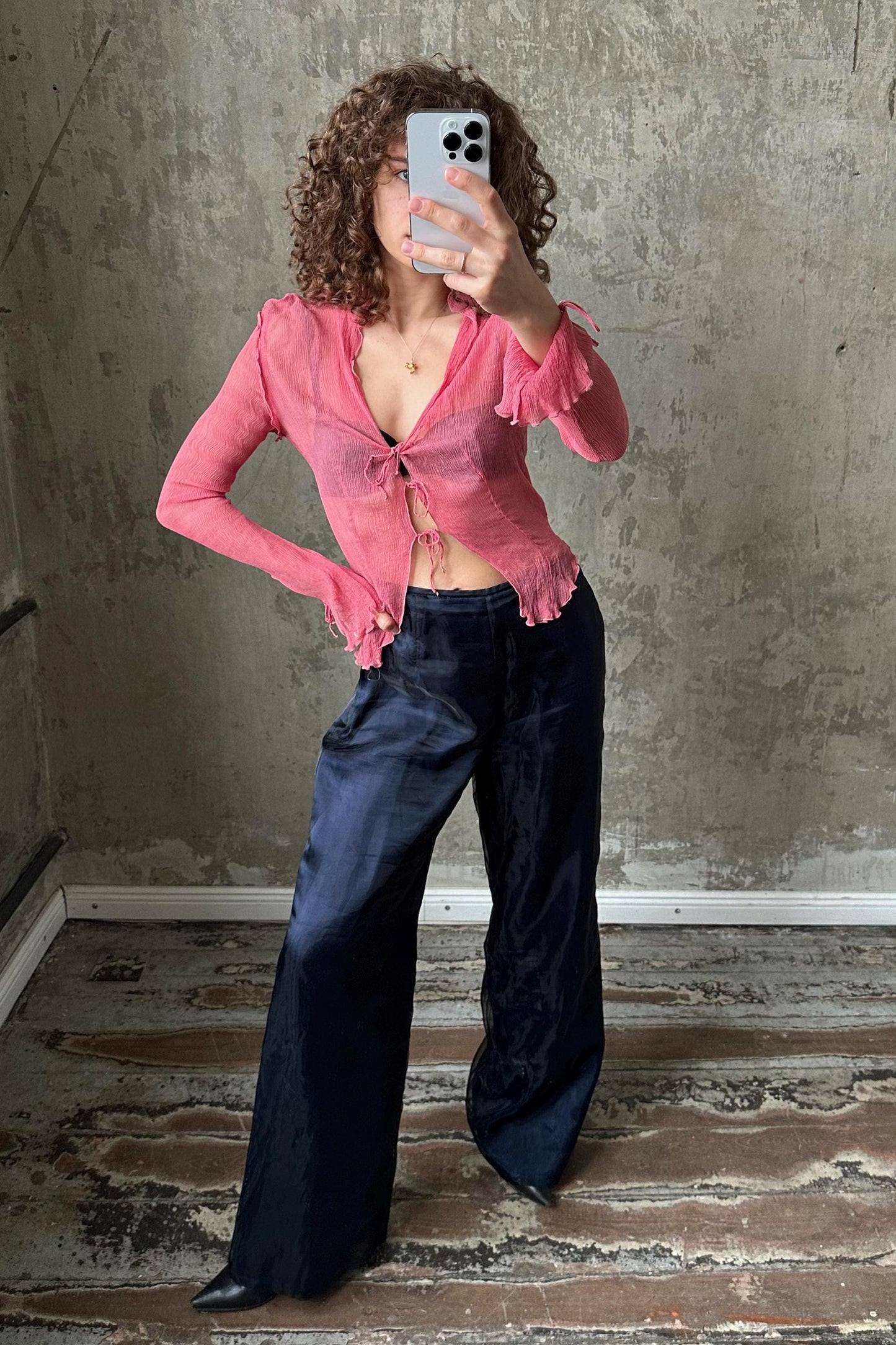 A woman with curly hair is taking a selfie with a smartphone, wearing a pink blouse and navy blue pants.