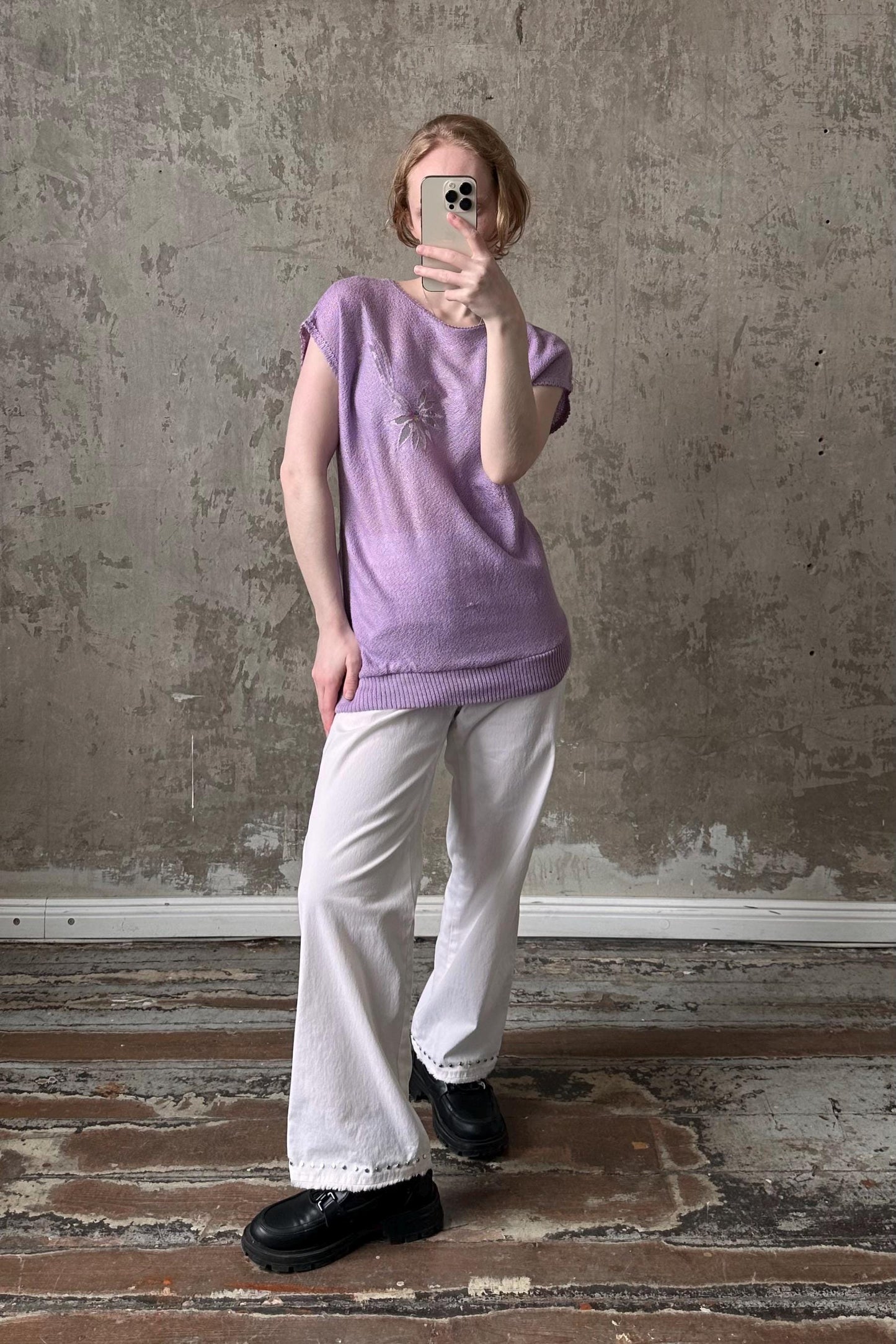 A person is taking a selfie with a smartphone, wearing a purple sweater and white pants, standing in front of a concrete wall.