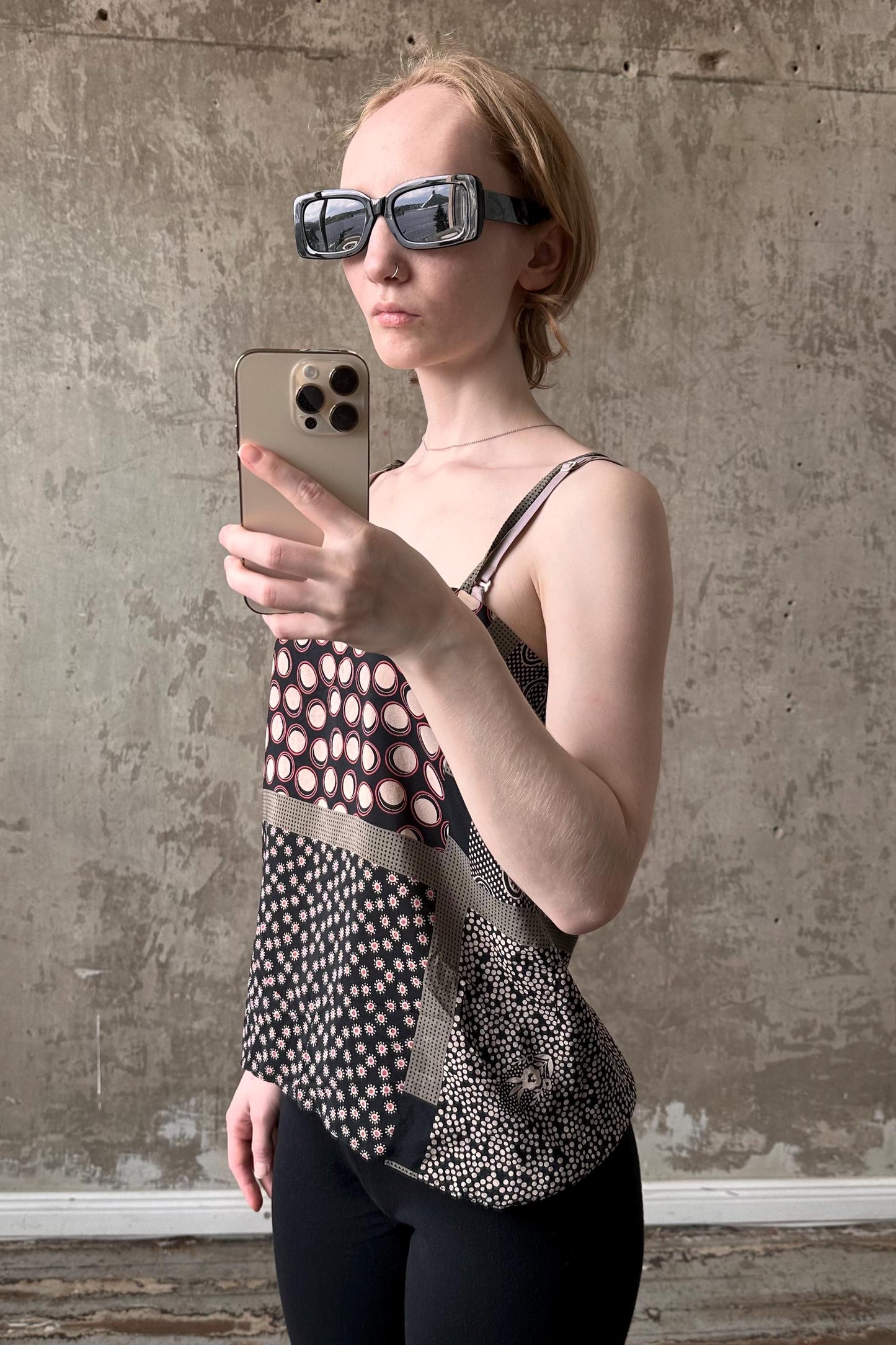 A woman wearing sunglasses and a patterned top is holding a smartphone and taking a selfie in front of a concrete wall.