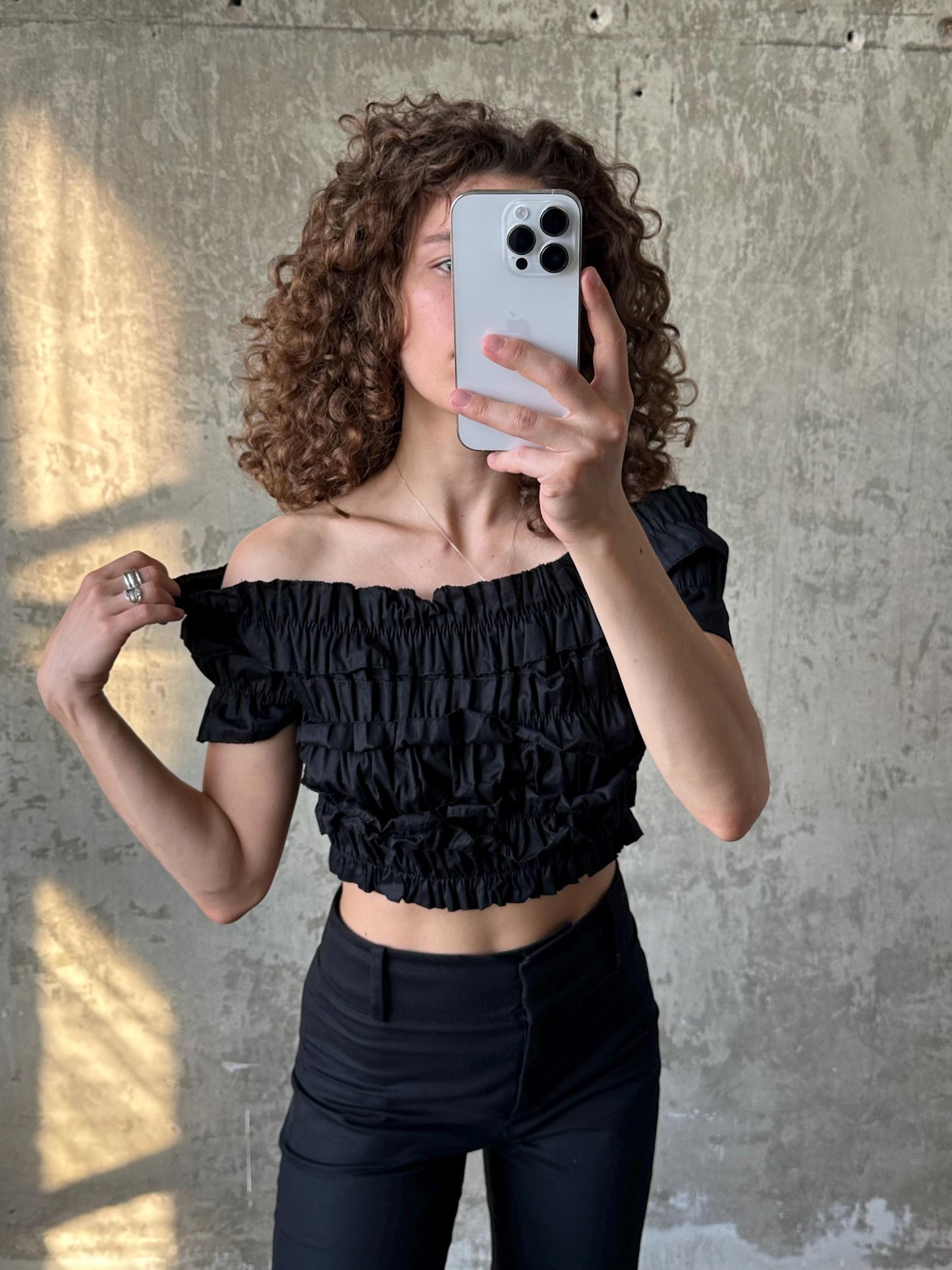 A woman with curly hair is taking a selfie with a smartphone, wearing a black off-the-shoulder top and black pants.