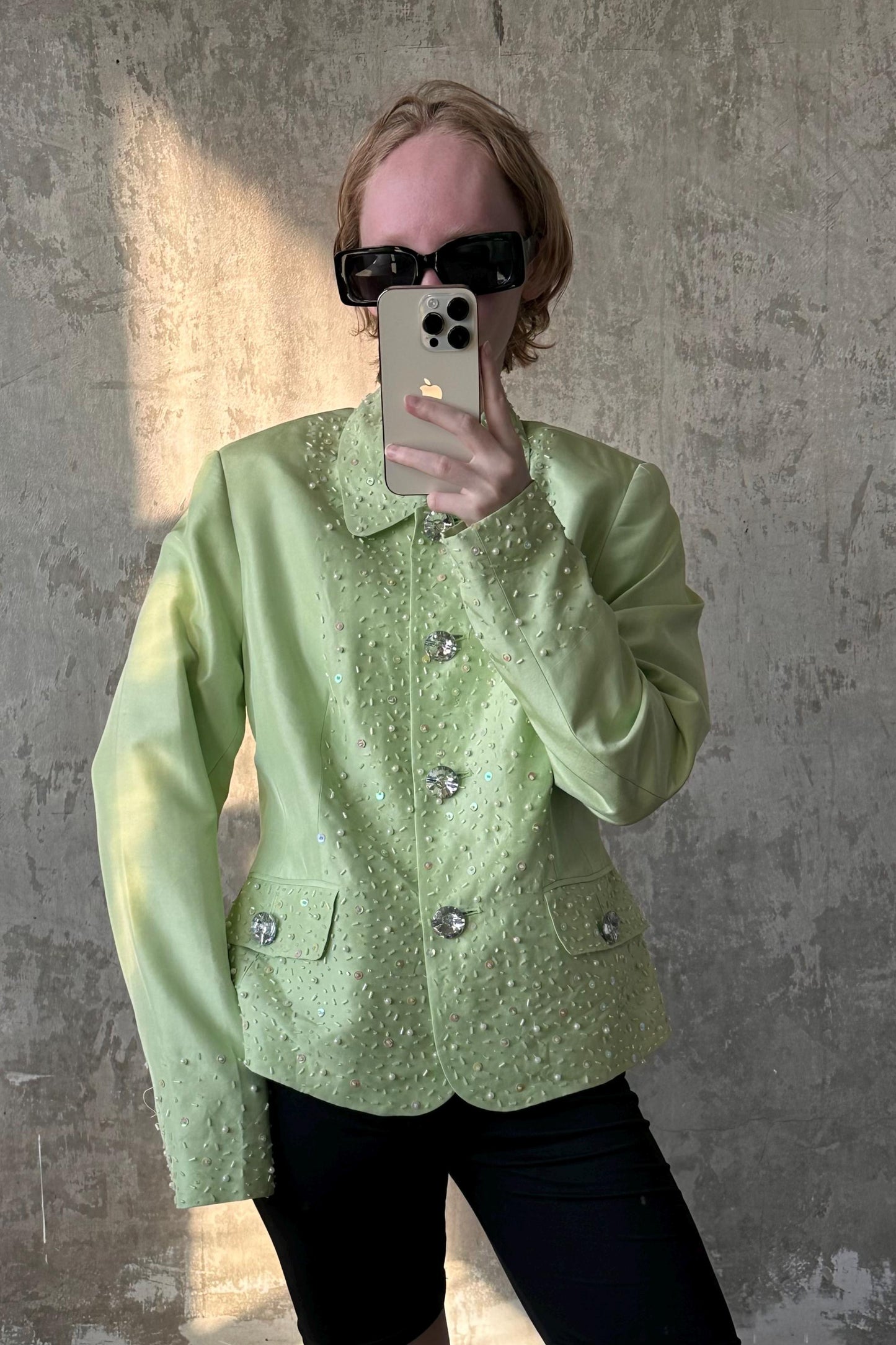 A person is taking a selfie with a green jacket and sunglasses, holding a smartphone in front of their face.