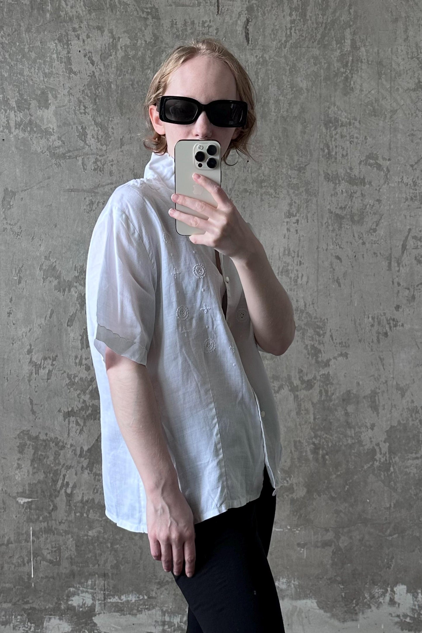 A person is taking a selfie with a white iPhone, wearing a white shirt and black pants, against a concrete wall background.