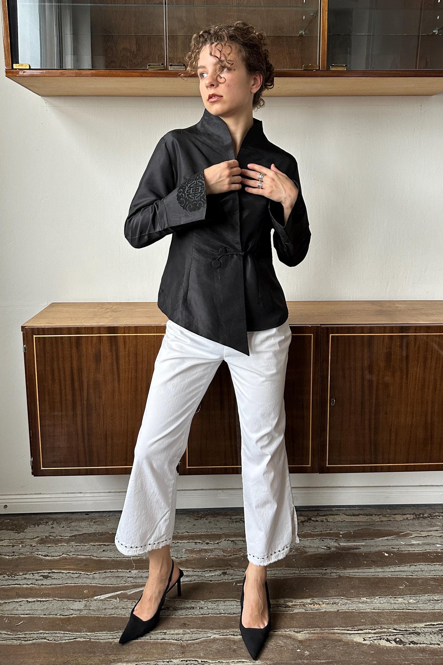 A woman wearing a black jacket and white pants stands in front of a wooden cabinet, adjusting her jacket.
