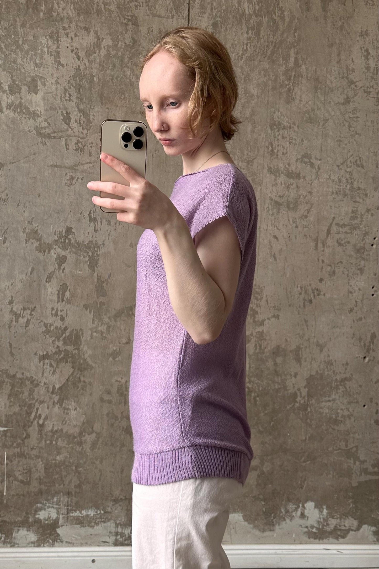 A woman in a purple sweater is taking a selfie with a smartphone, standing in front of a concrete wall.