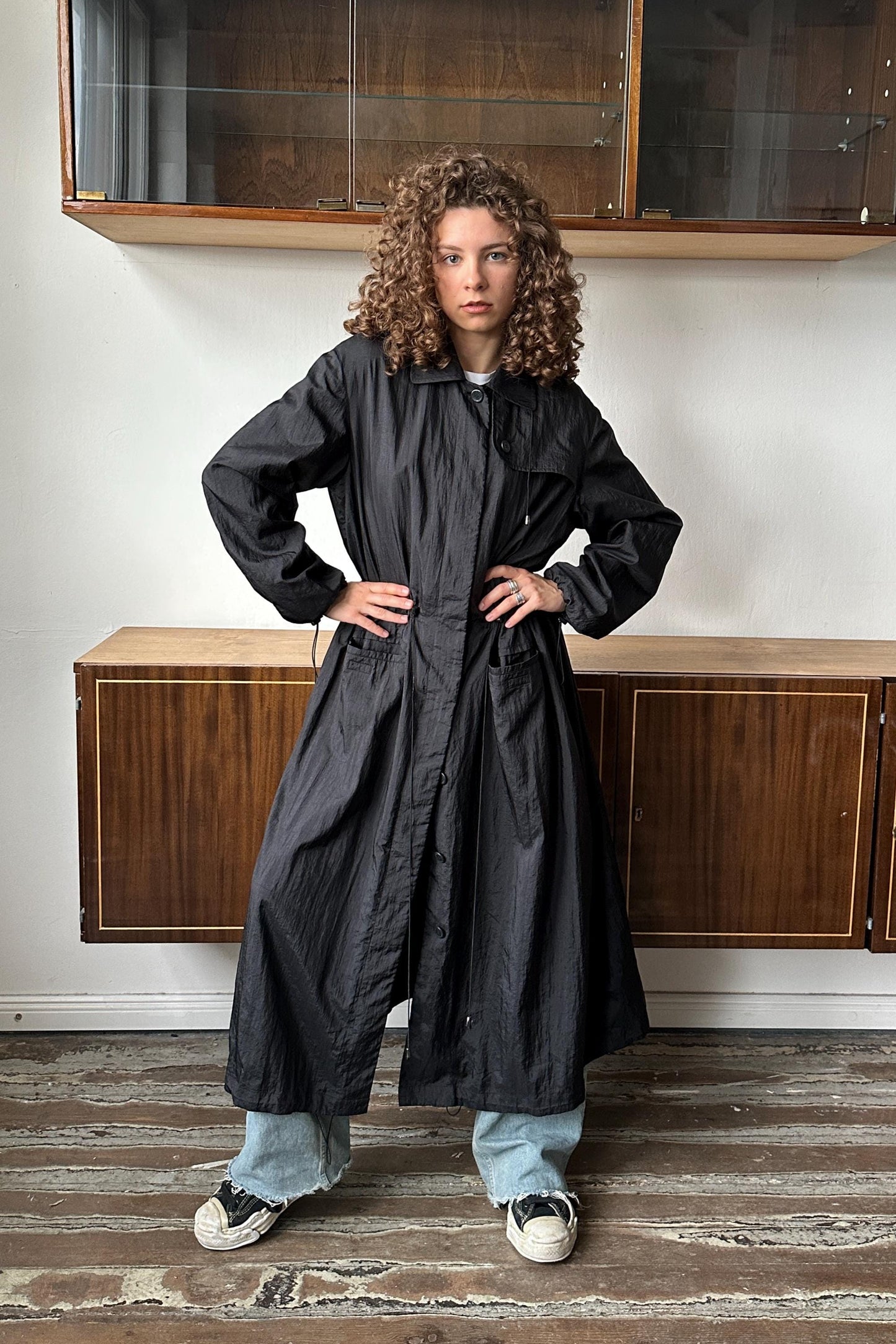 A young woman with curly hair stands confidently in a room with wooden cabinets and a striped floor.