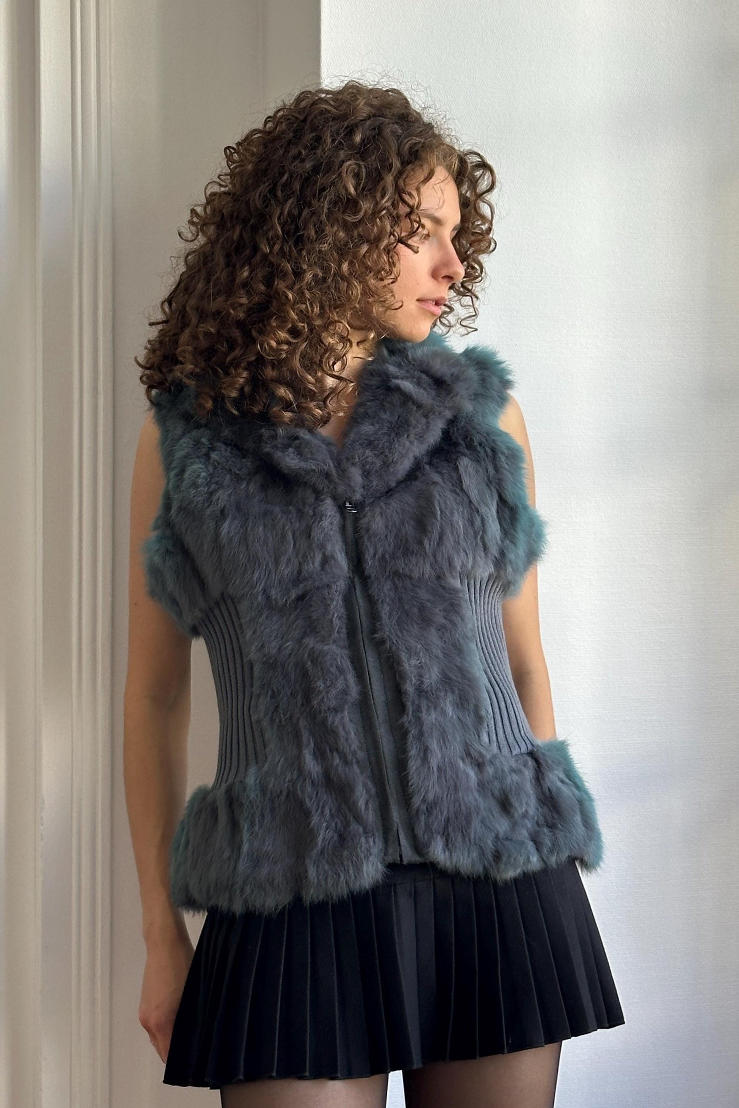 A woman with curly hair wearing a gray fur vest and a black skirt stands in front of a white wall.