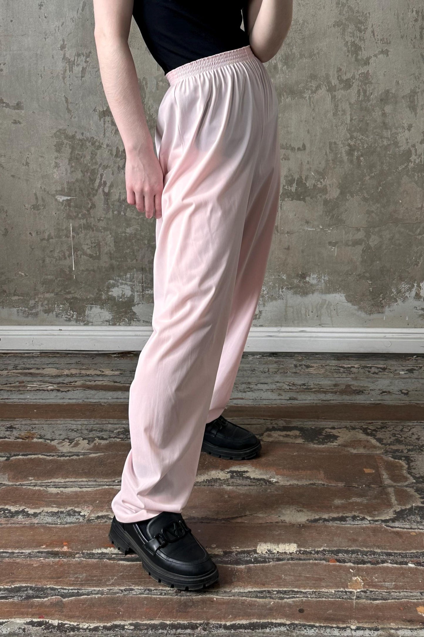 A person wearing a black top and pink pants stands in front of a concrete wall, with their hands in their pockets.