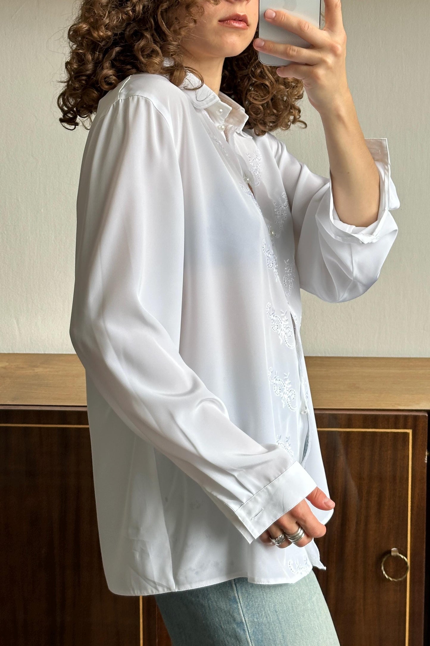 A woman with curly hair is taking a selfie with a white blouse and a wooden cabinet in the background.