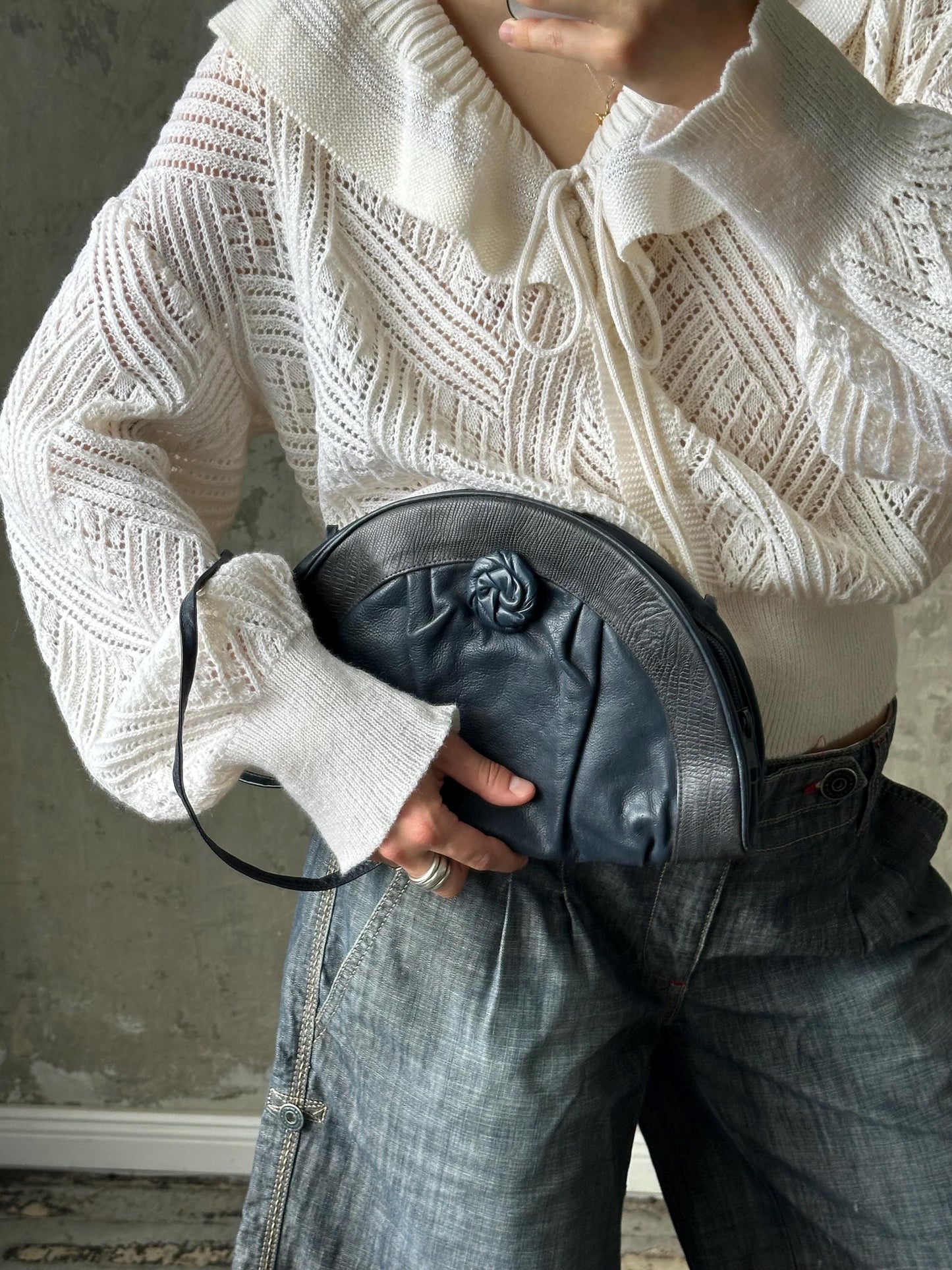 A person wearing a white sweater and blue jeans is holding a black purse with a ruched design.
