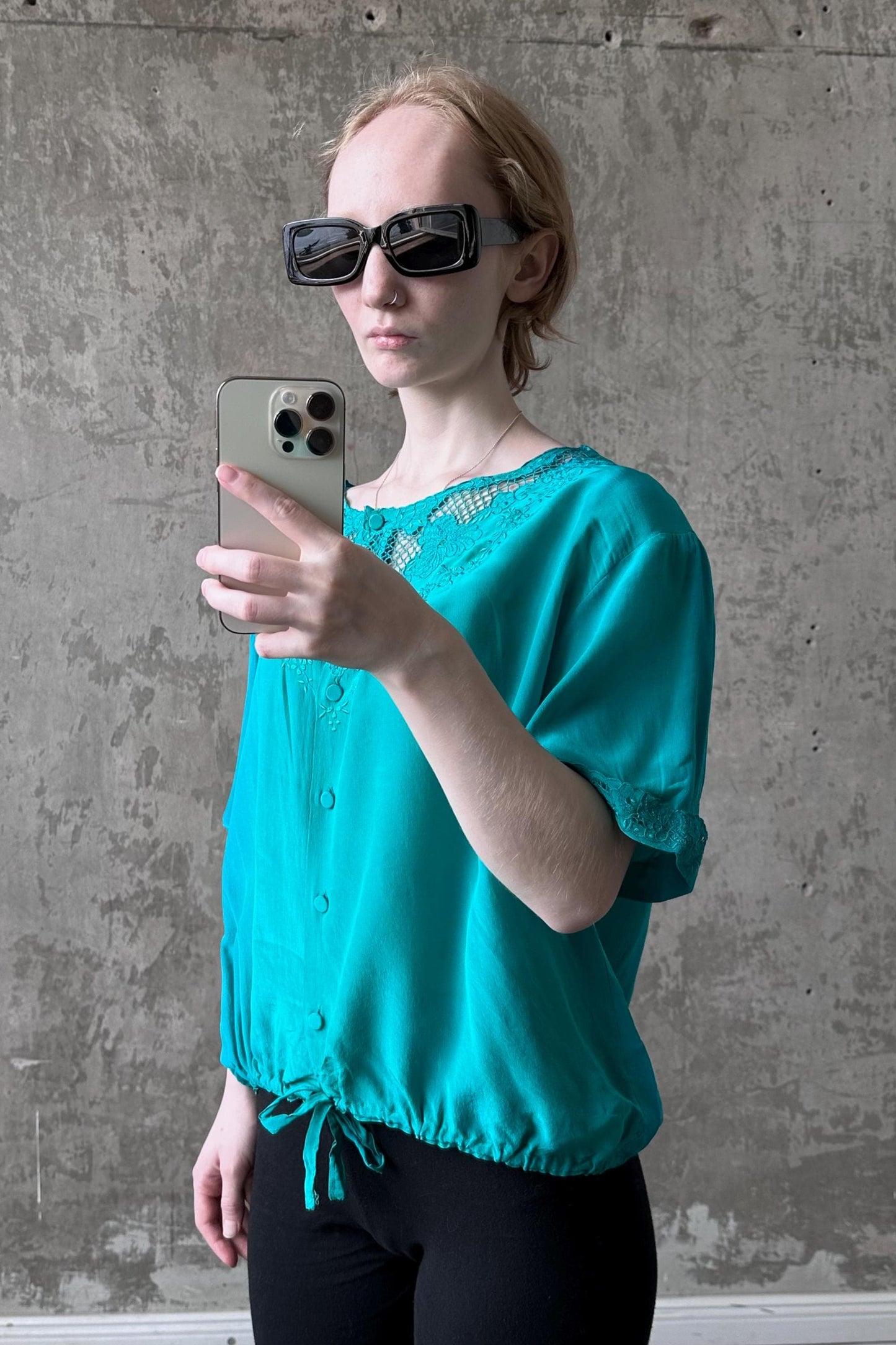 A woman wearing a turquoise blouse and sunglasses is taking a selfie with a smartphone.