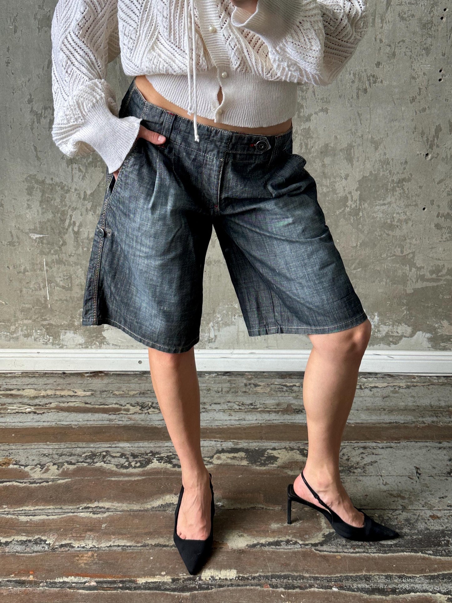 A person wearing a white sweater, blue jeans, and black heels stands in front of a concrete wall.