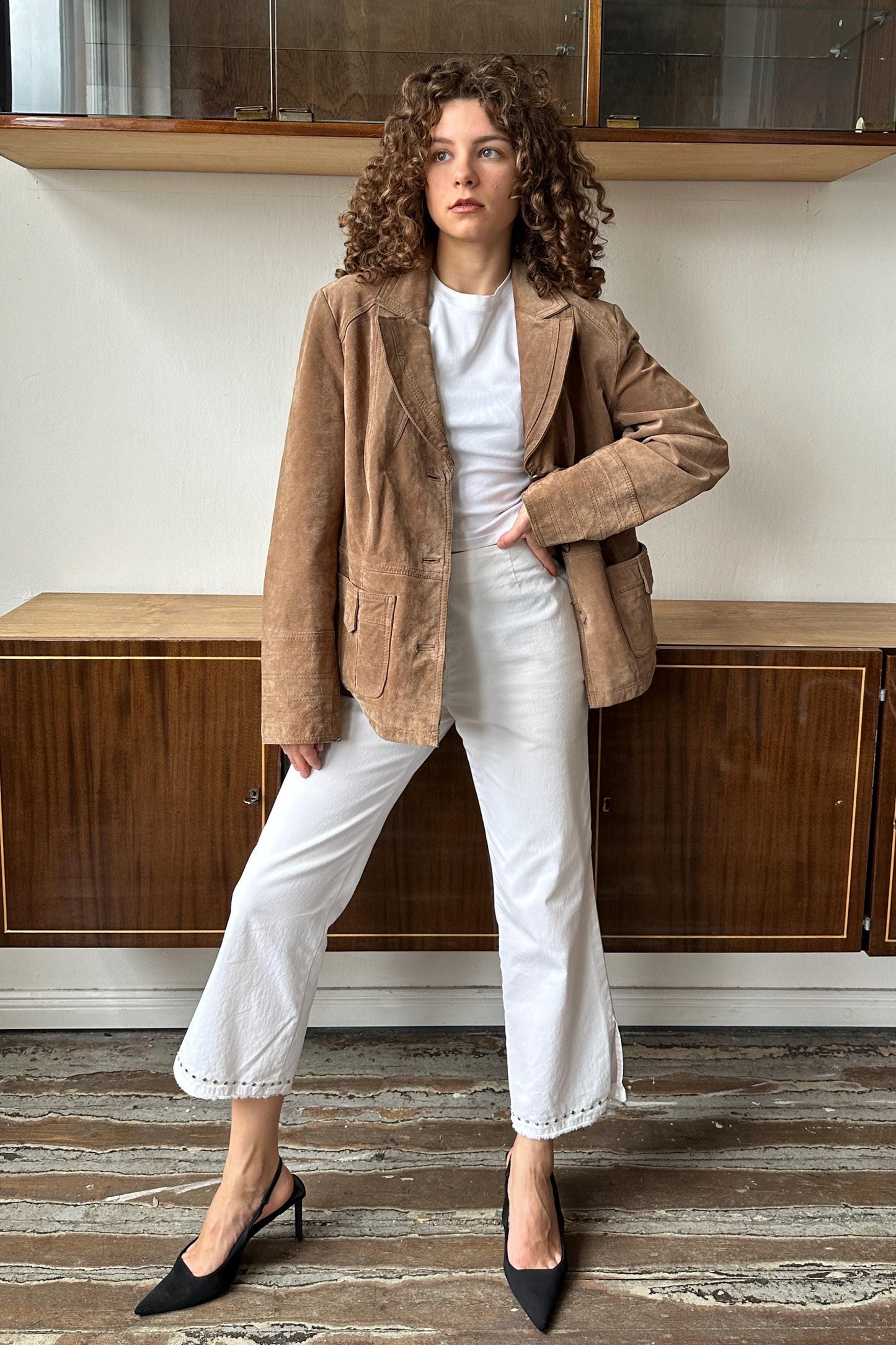 A woman with curly hair wearing a brown jacket, white pants, and black heels stands in front of a wooden cabinet.