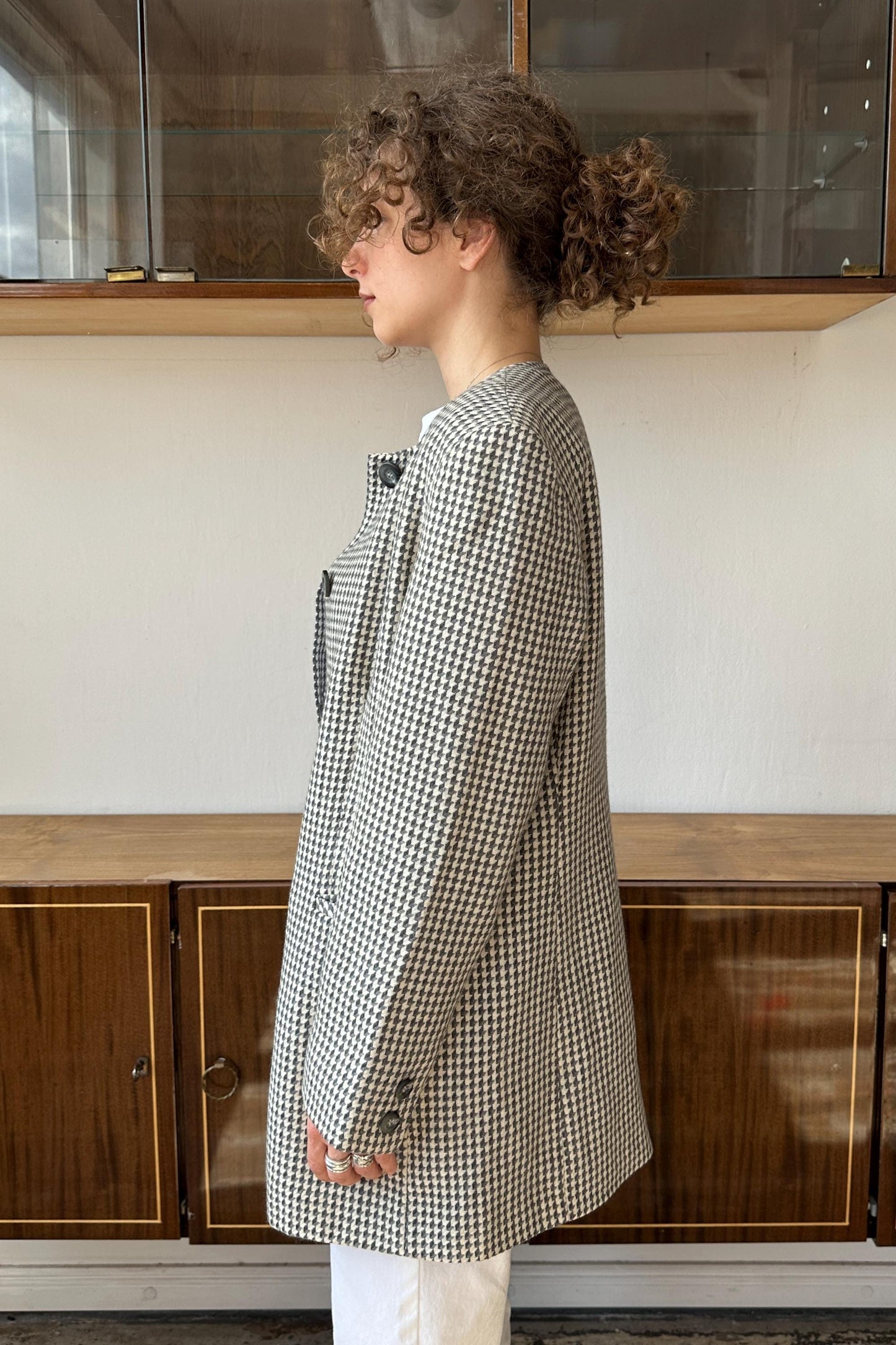 A woman with curly hair wearing a black and white checkered jacket stands in front of a wooden cabinet.
