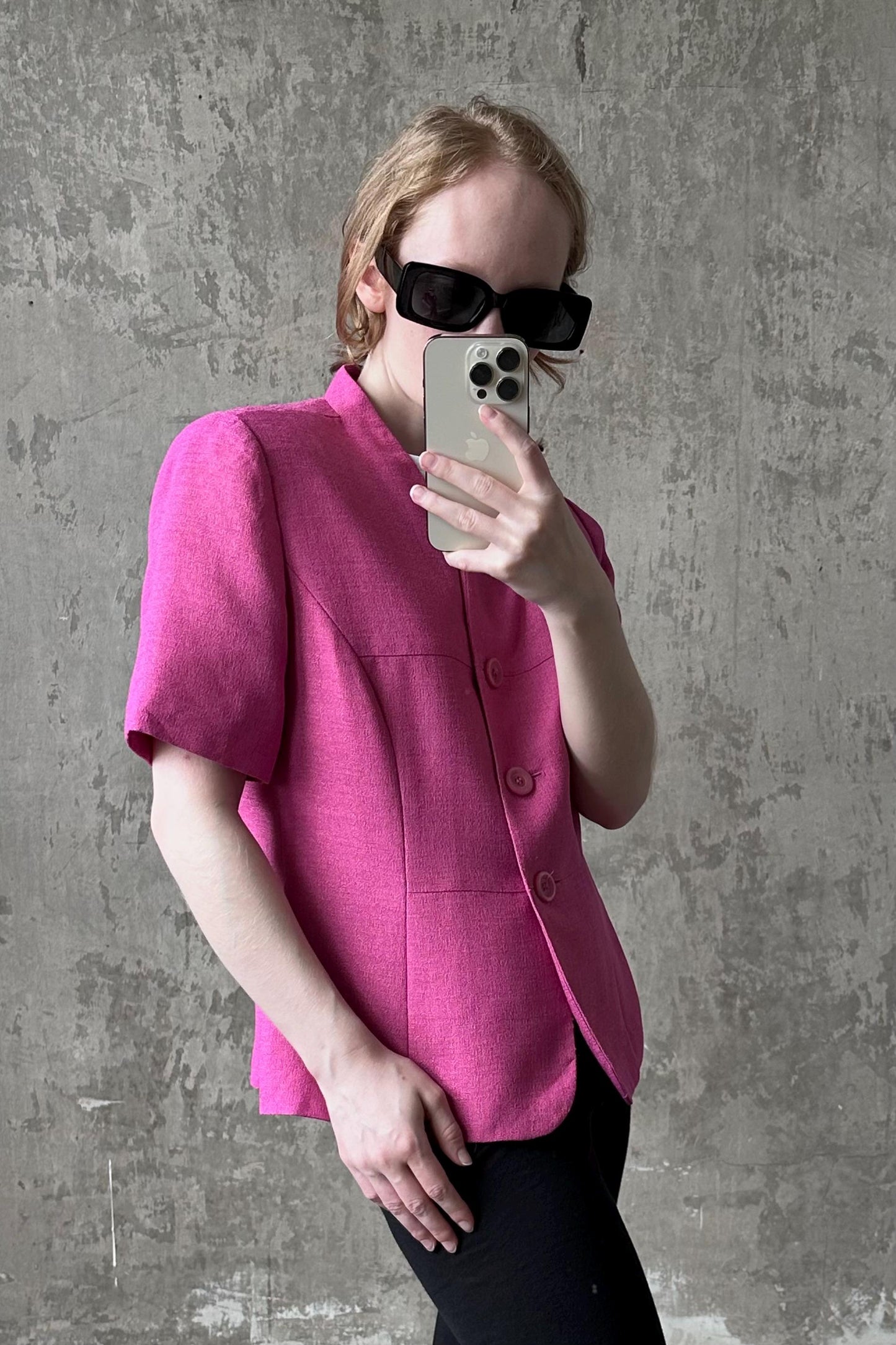 A woman wearing a pink blazer and sunglasses is taking a selfie with her iPhone against a concrete wall.