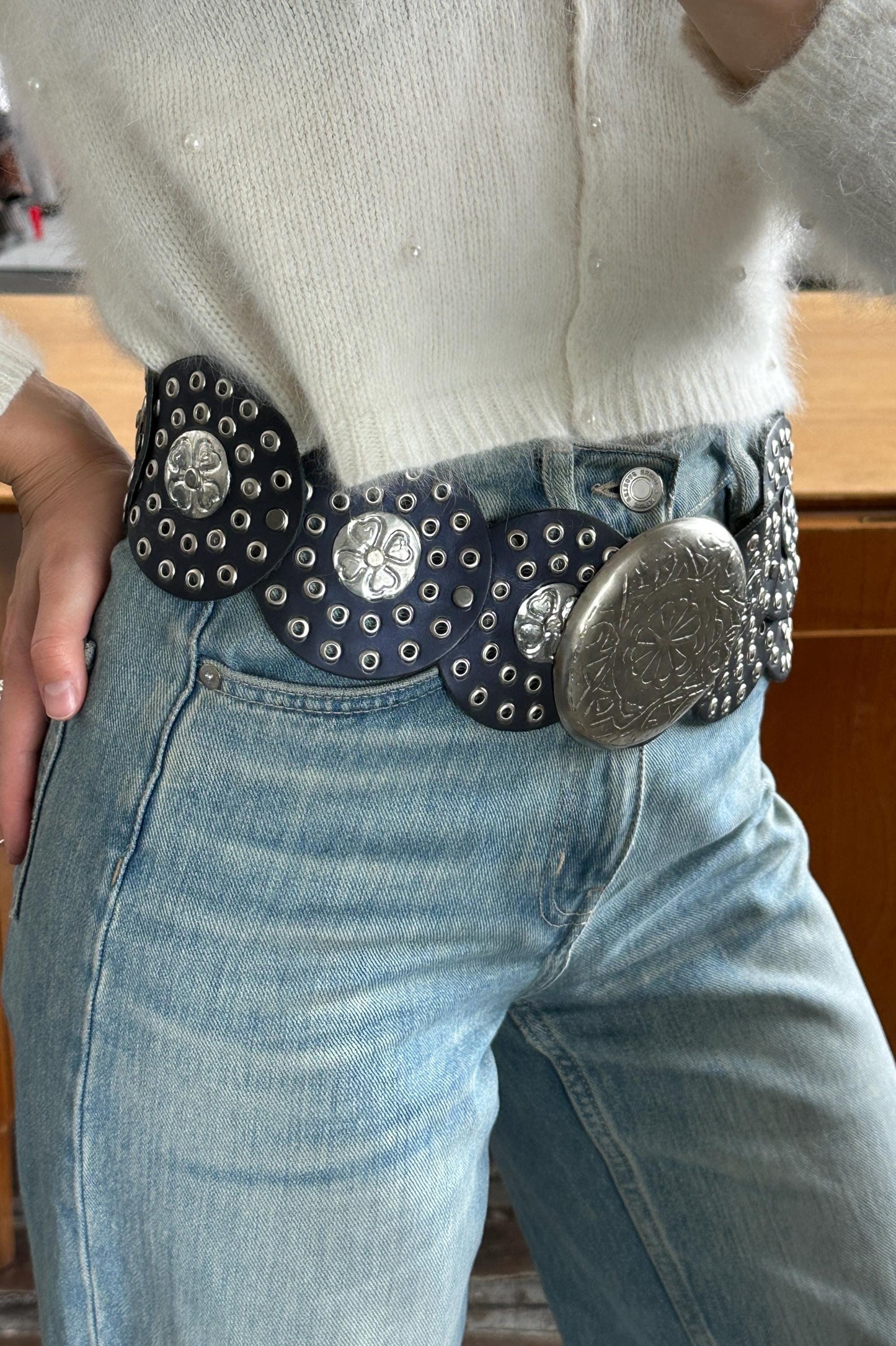 A person wearing a white sweater and blue jeans, with a large, decorative belt featuring metal studs and a metal buckle.