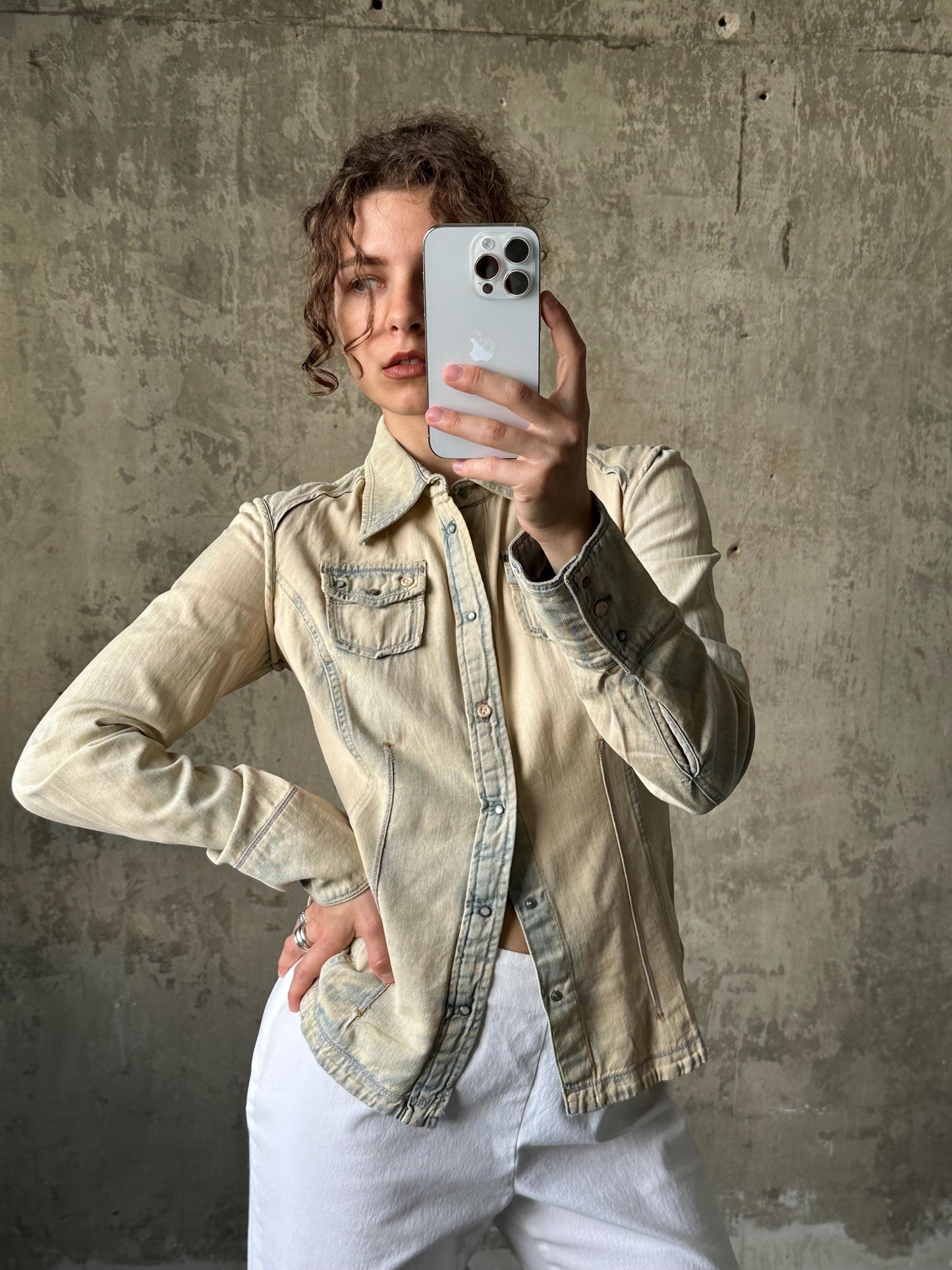 A person is taking a selfie with a smartphone, wearing a beige denim jacket and white pants, against a concrete wall background.