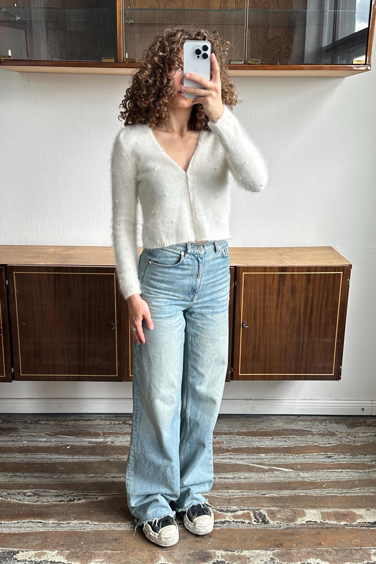 A woman with curly hair is taking a selfie with her phone, standing in front of a wooden cabinet.